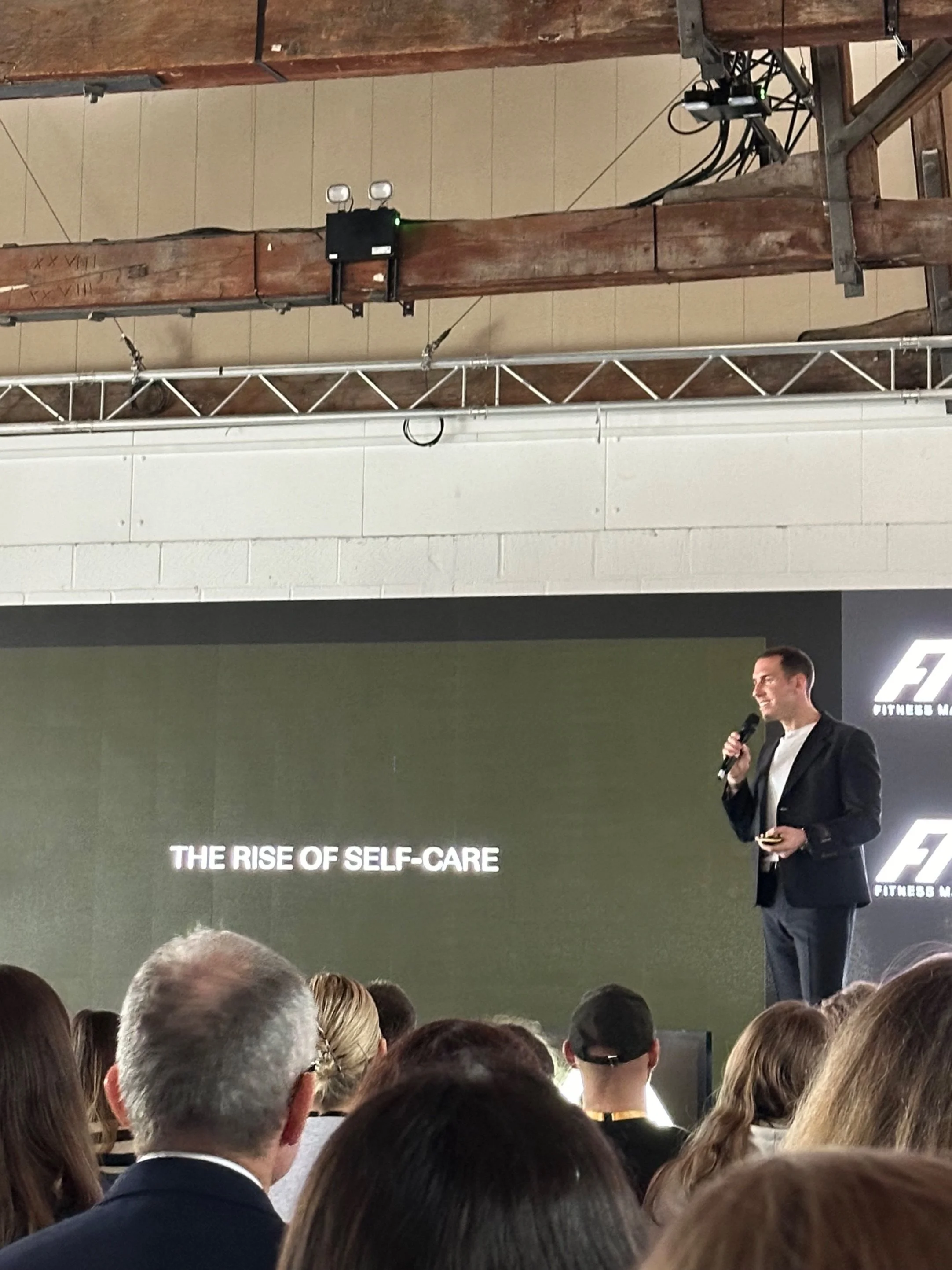 Speaker on stage presenting "The Rise of Self-Care" to an audience; equipment visible on ceiling.
