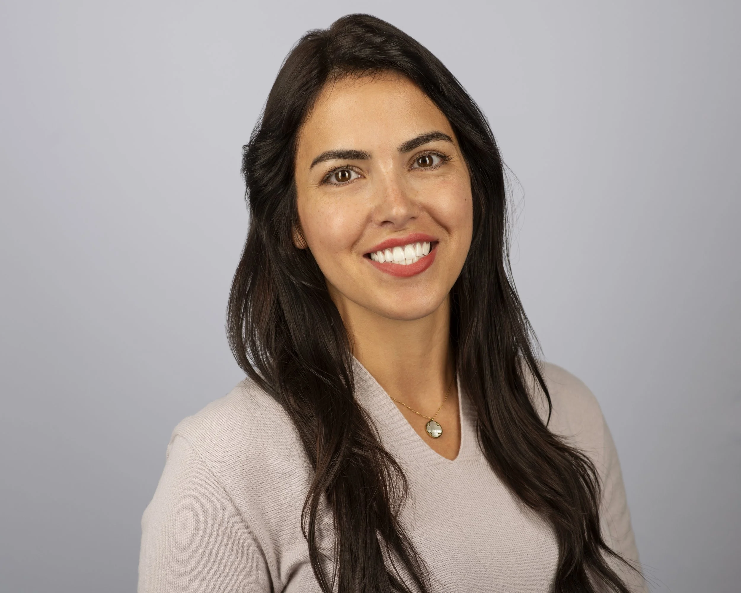 Smiling woman with long dark hair wearing a necklace and light sweater against a neutral background.