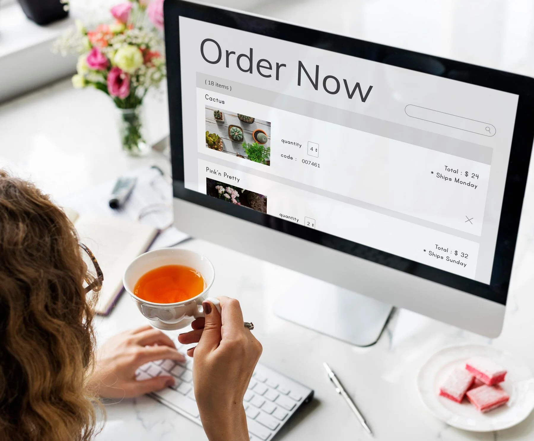 A woman holding a cup of tea while using a desktop computer to browse an online store. The screen displays an order summary with plant items listed. There's a notebook, pen, and sweets on the desk, with flowers in the background.