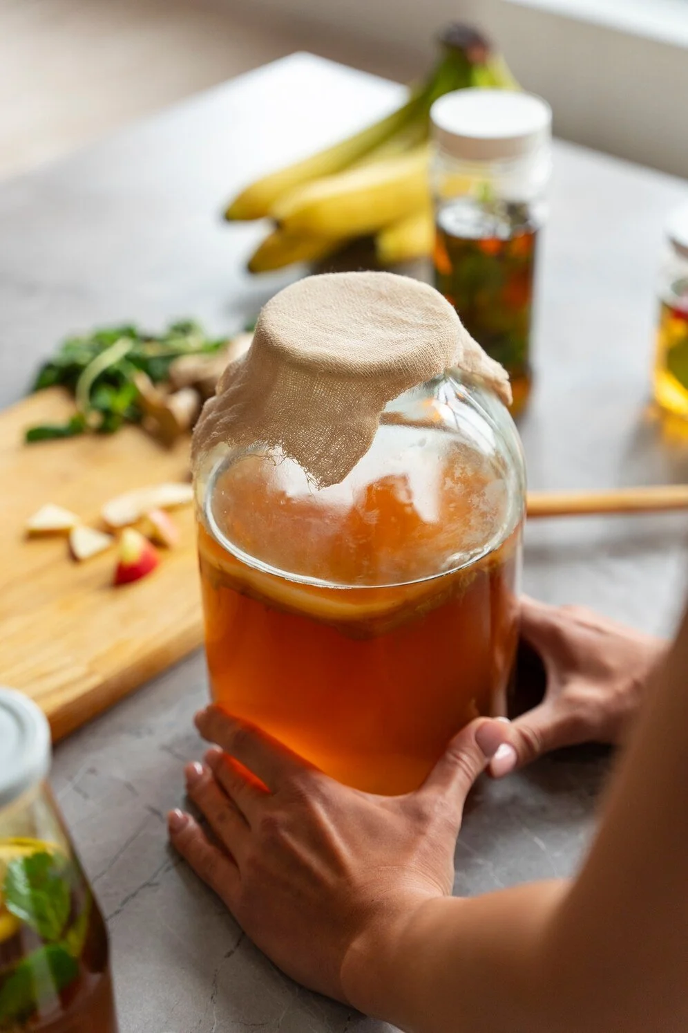 Person holding a jar of kombucha with cloth cover, bananas and herbs on a table
