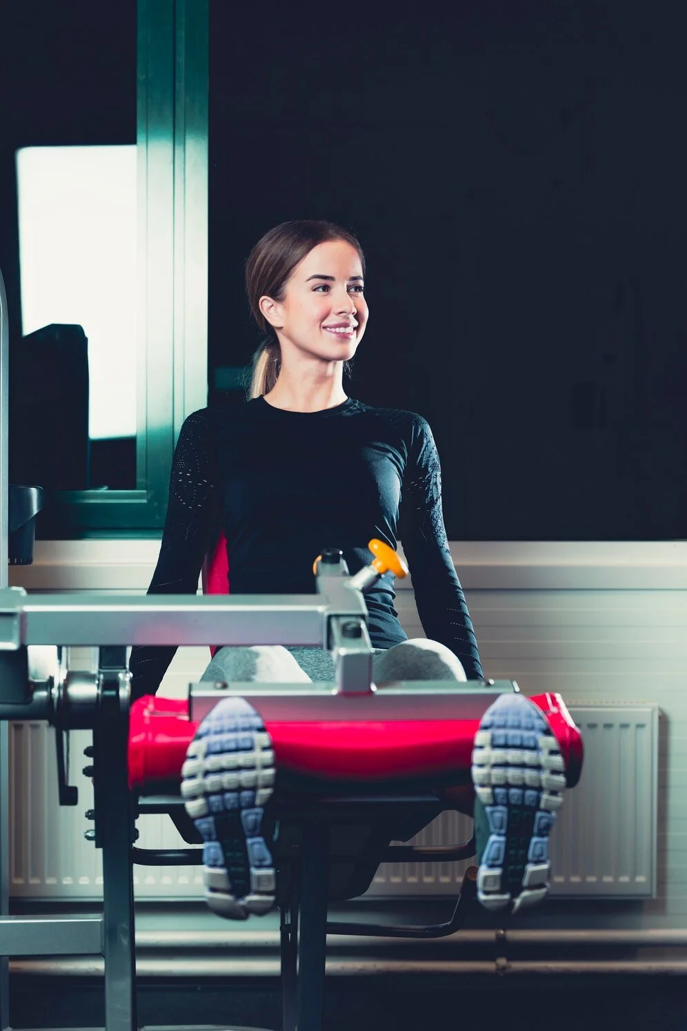 Woman exercising on a leg extension machine in a gym, wearing a black long-sleeve shirt and athletic pants, smiling.