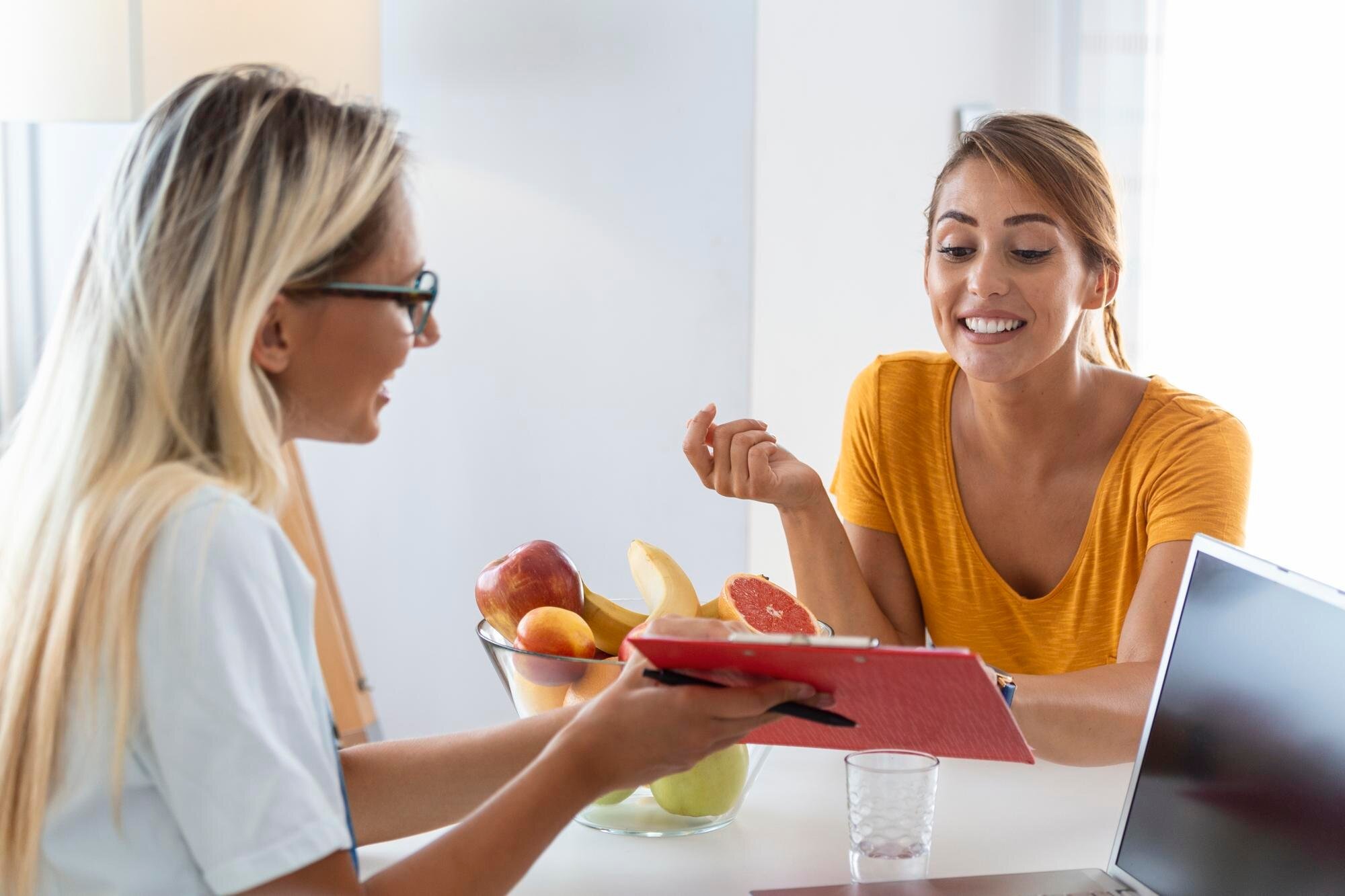 Two women discussing at a table with a laptop and a bowl of fruit, one holding a red notebook, the other smiling.