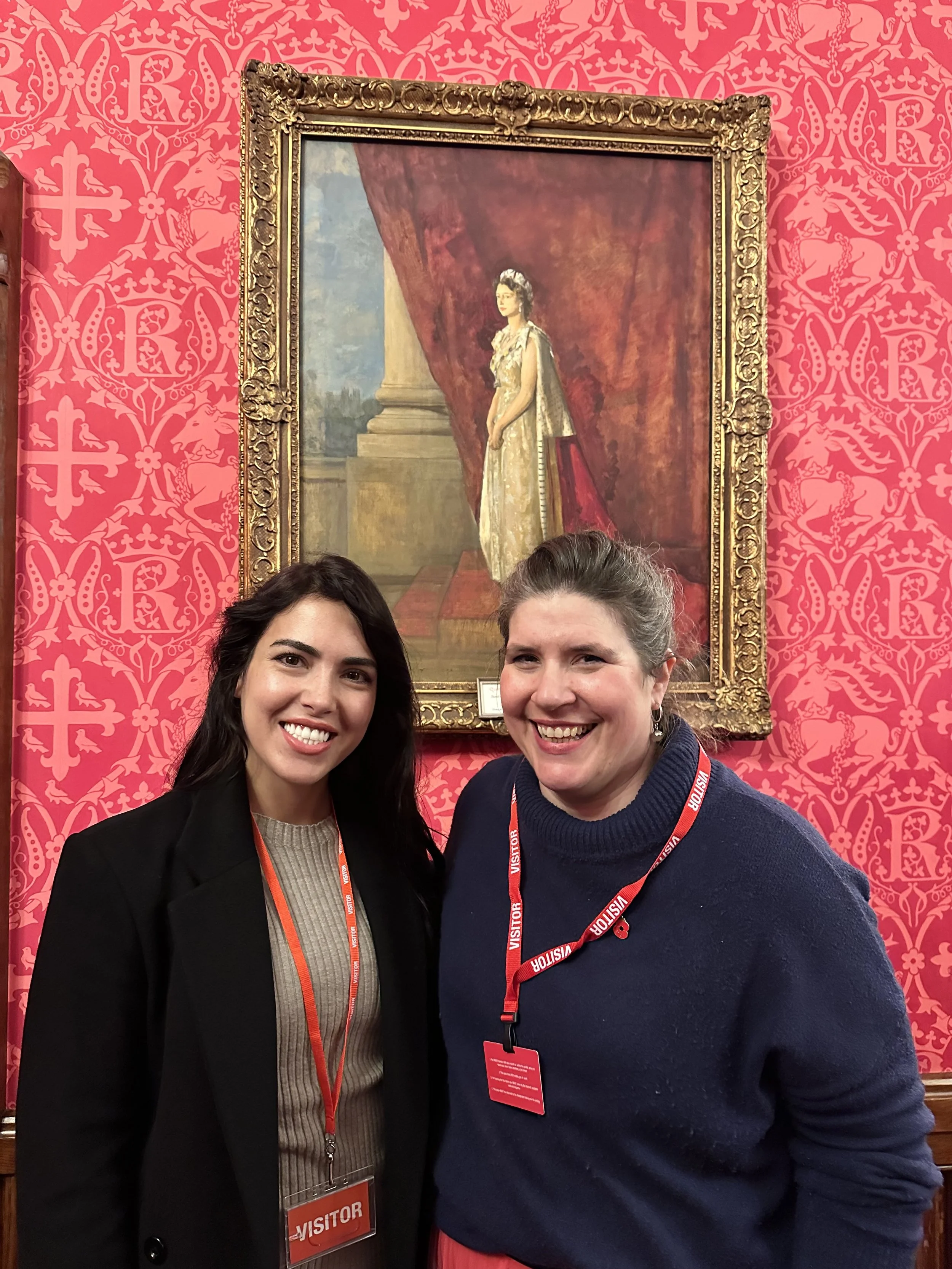 Two women smiling in front of a framed portrait in a room with ornate red wallpaper. Both are wearing visitor badges.
