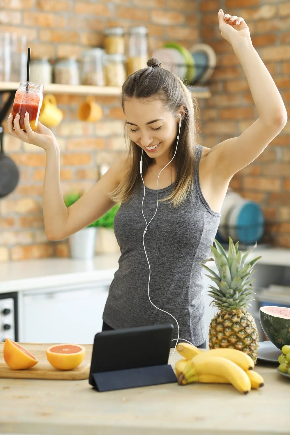 Woman in a kitchen enjoying music with headphones, holding a smoothie, surrounded by fresh fruits like pineapple, banana, and grapefruit, with a tablet on the counter.