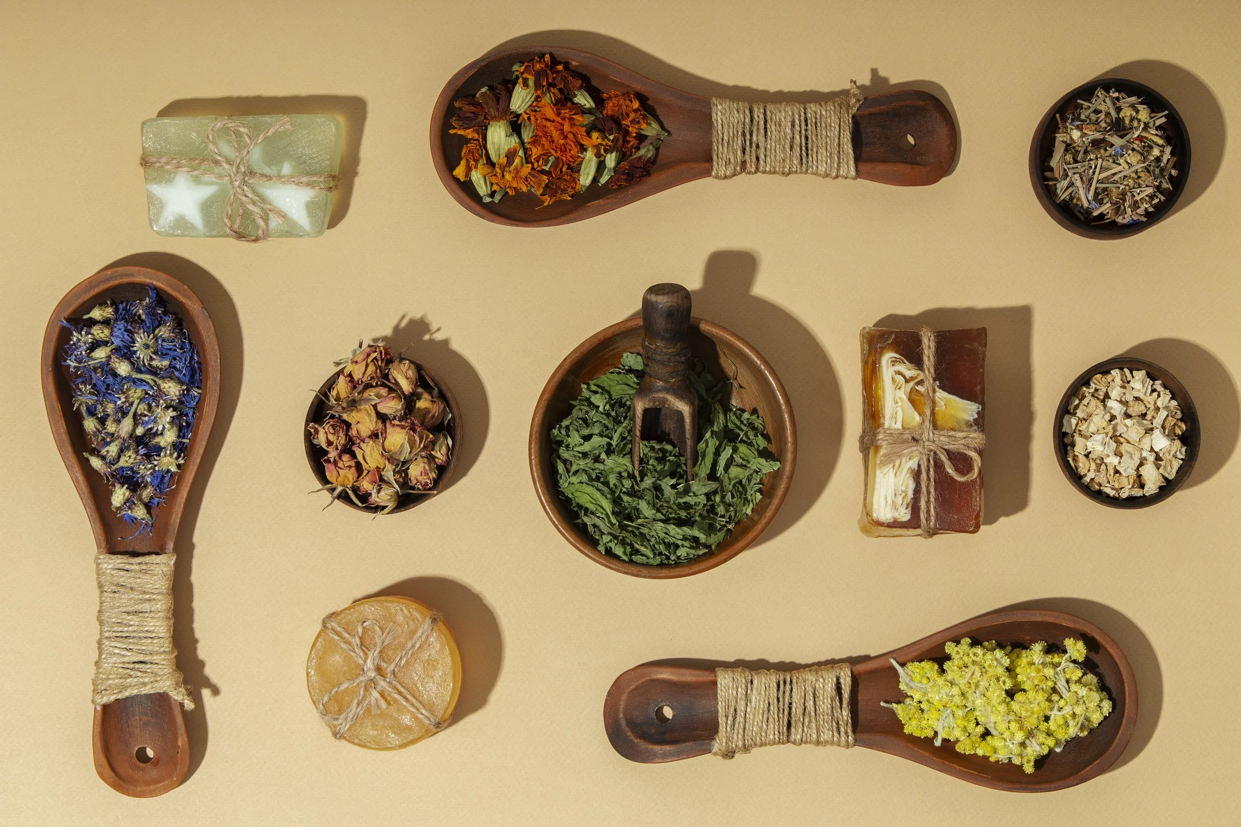 An arrangement of various dried herbs, flowers, and two bars of soap on a beige background. Includes wooden spoons and bowls holding different botanical ingredients such as blue and yellow flowers, rose buds, and leafy greens.