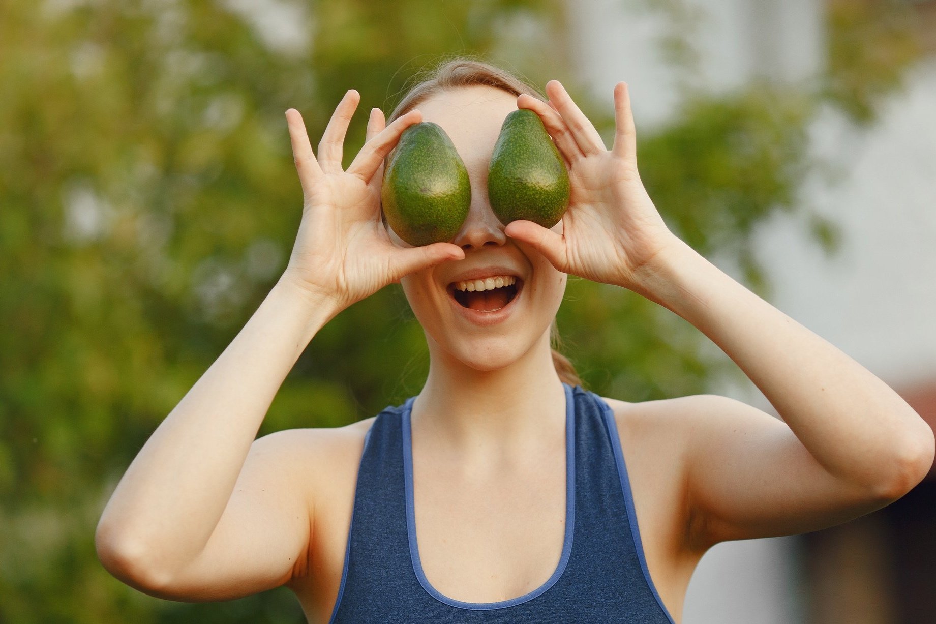 Woman holding avocados in front of eyes, smiling, outdoor setting.
