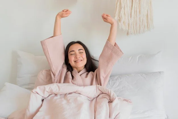 Woman stretching in bed with a relaxed expression, wearing a pink robe, in a cozy room setting.