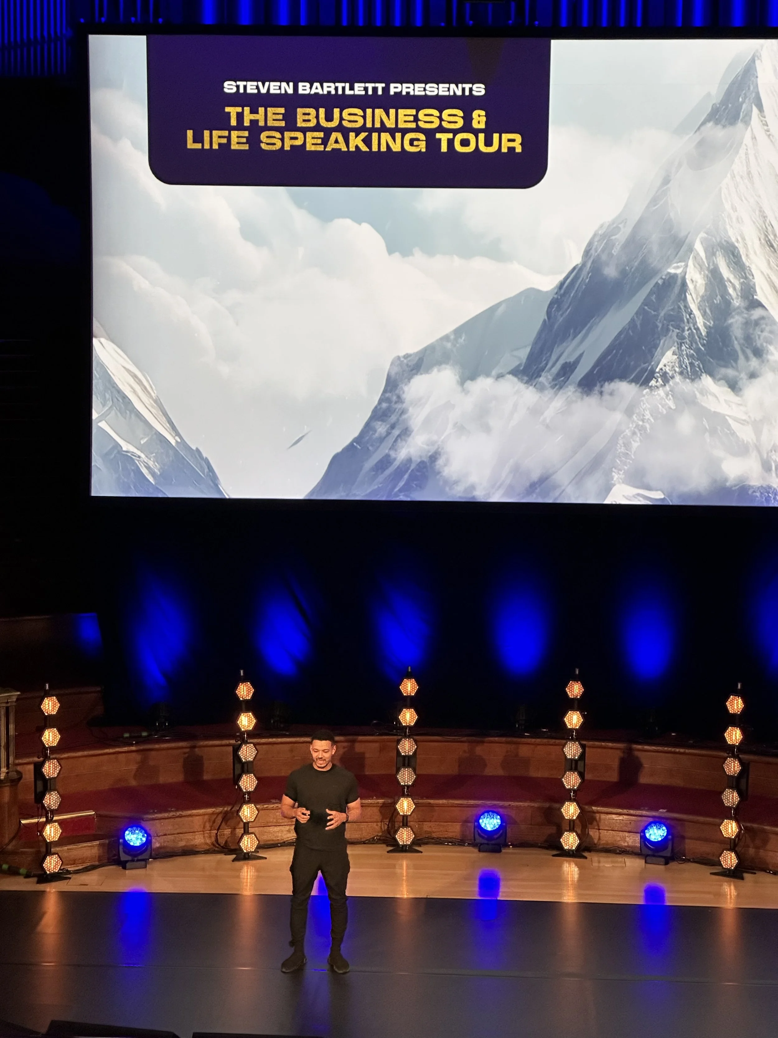 A person on stage in front of a large screen displaying 'Steven Bartlett Presents The Business & Life Speaking Tour' with a mountain background. The stage is illuminated with blue lighting and decorative light stands.