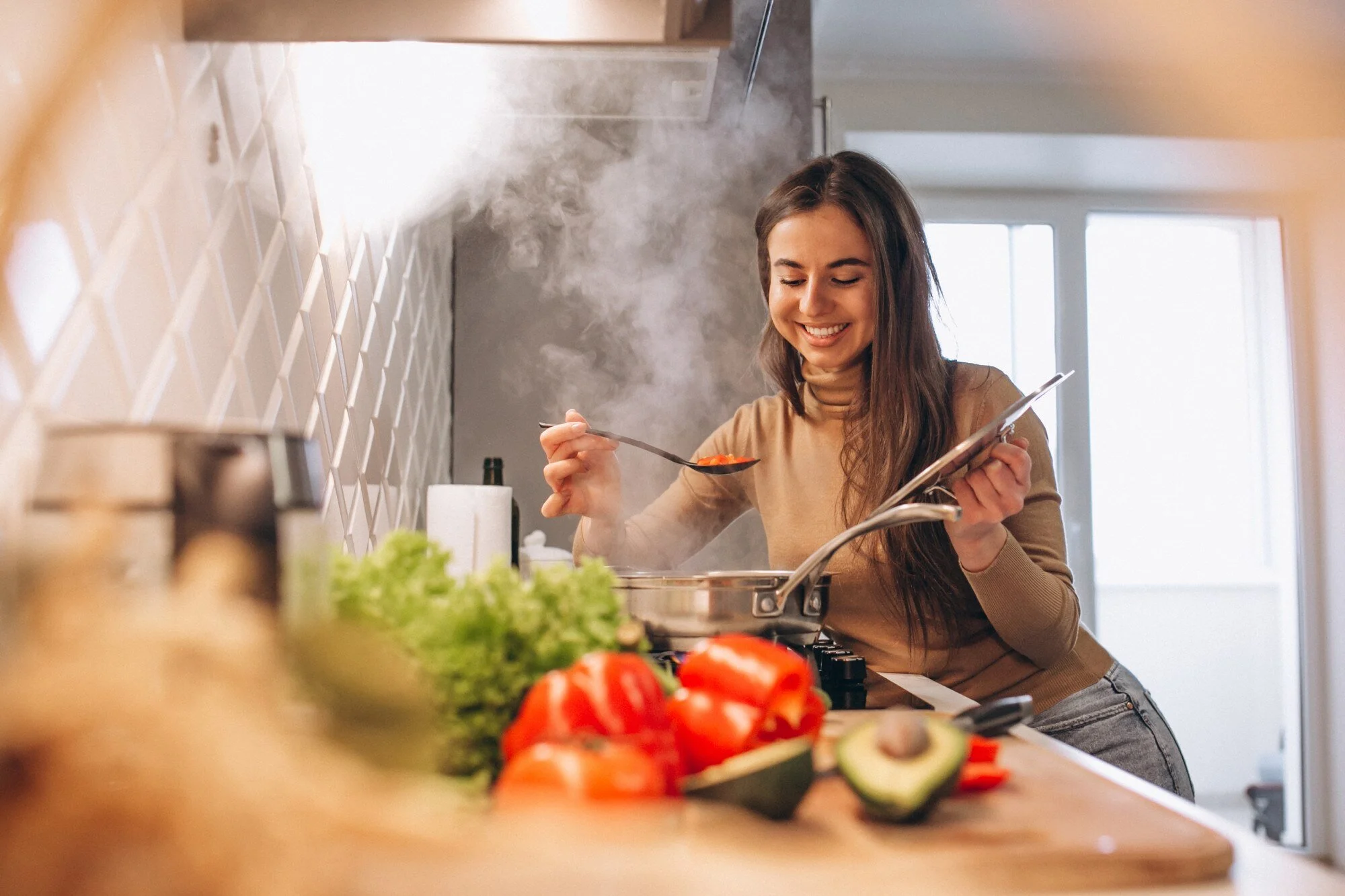 Woman cooking in a kitchen, tasting food with spoon over steaming pot, surrounded by fresh vegetables like bell peppers and avocado.