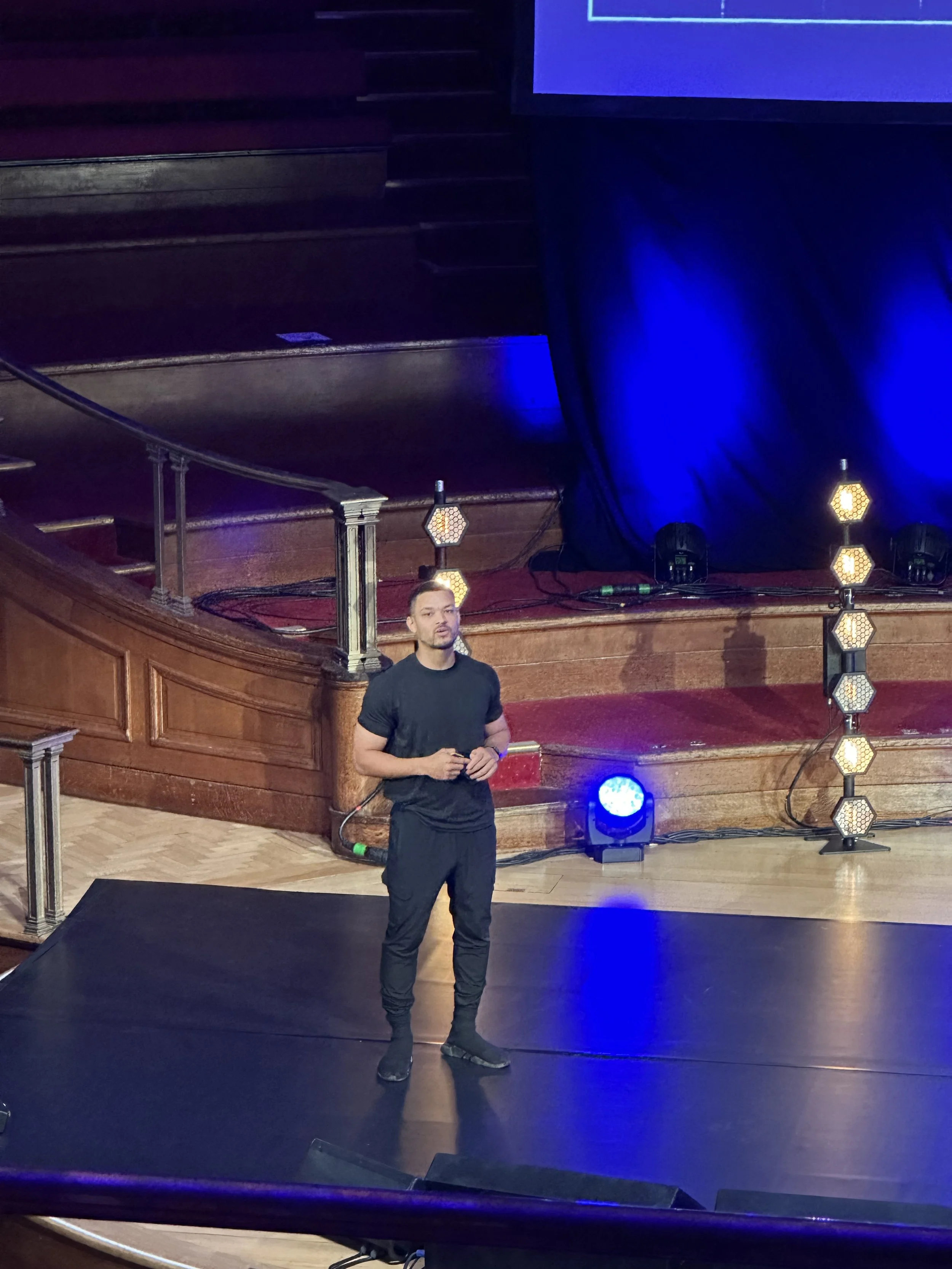 Person standing on stage with black shirt and pants, under blue lighting.