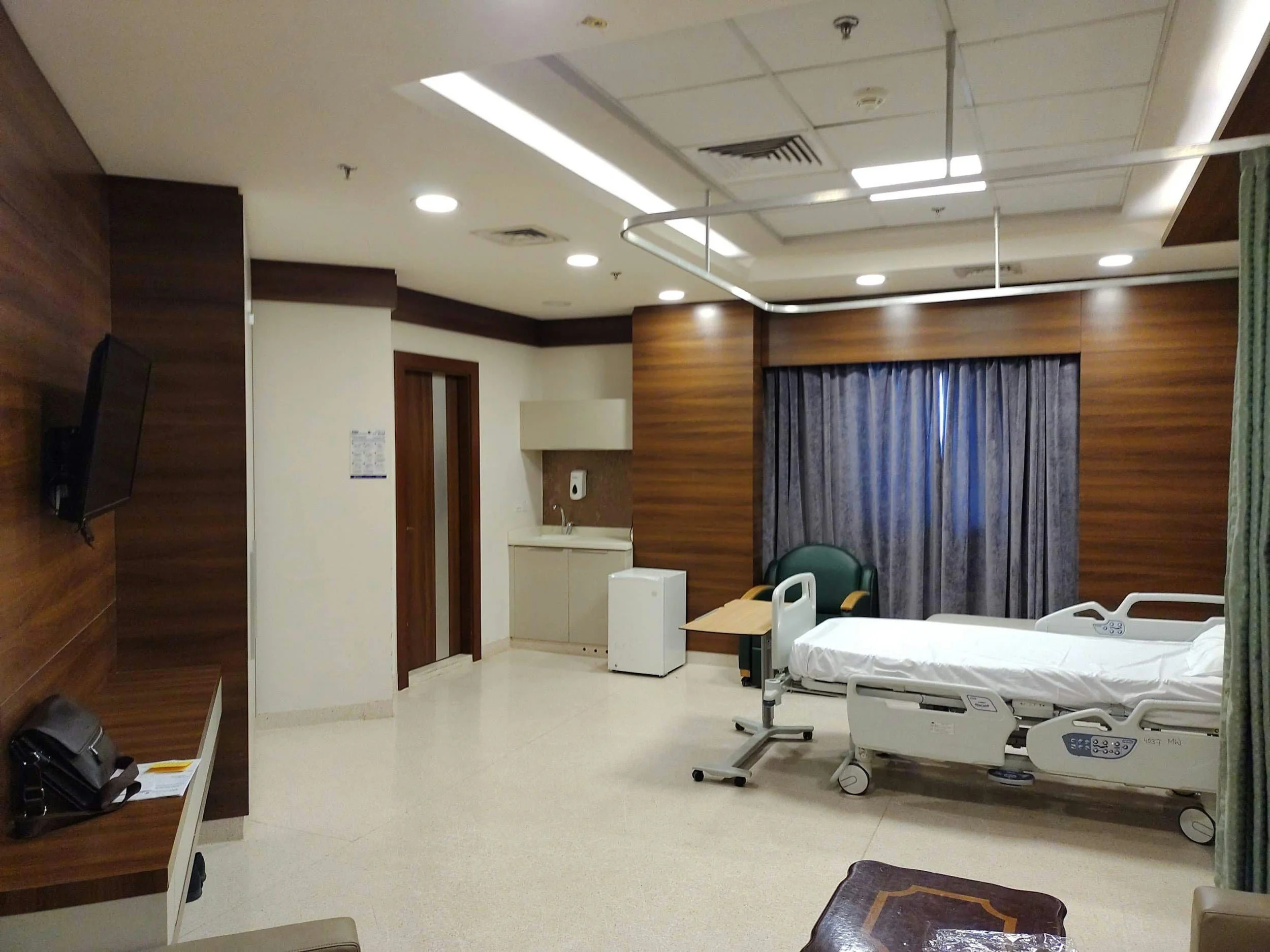 A modern hospital room with a single bed, medical equipment, a chair, TV, and wooden paneling on the walls.