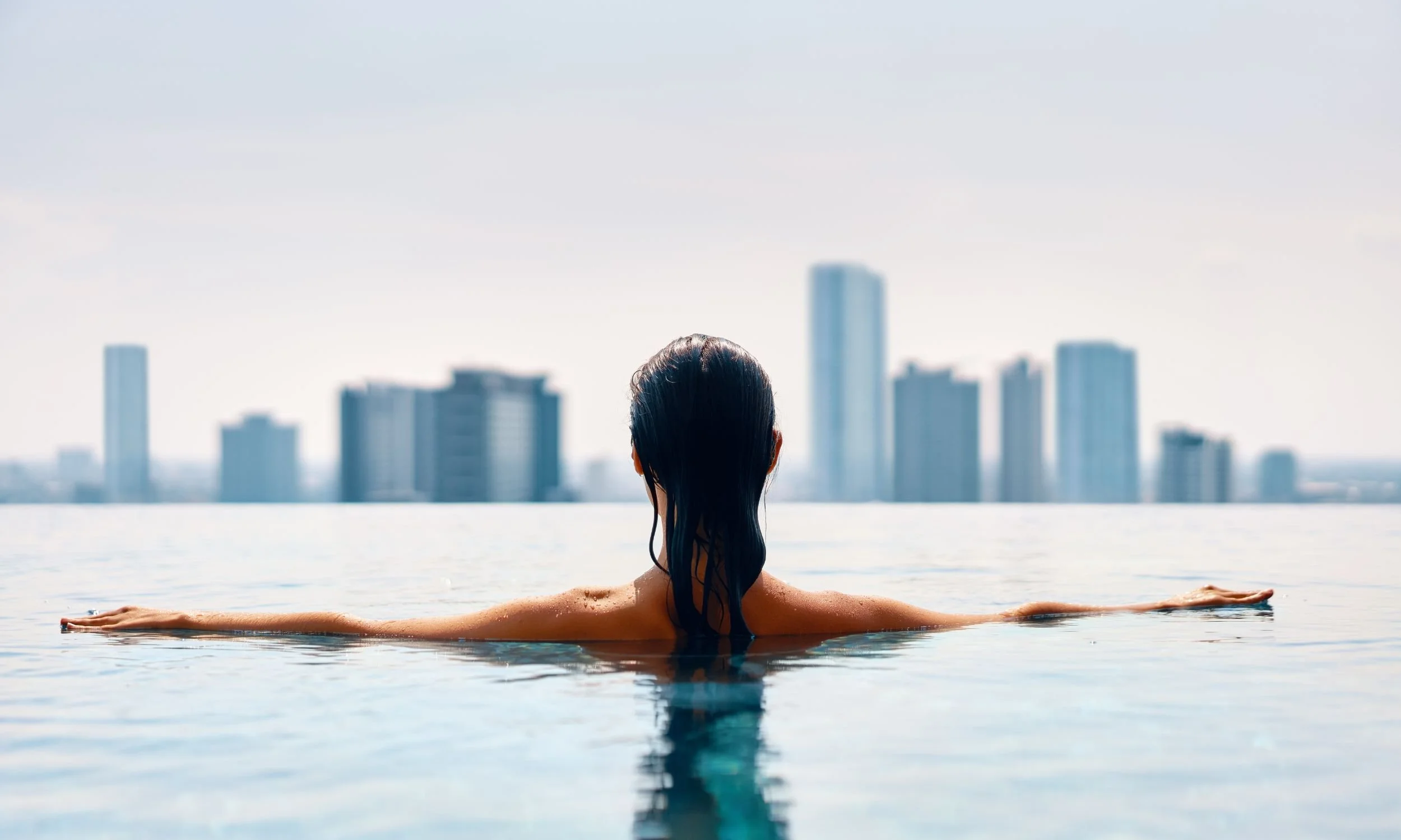 A woman in a rooftop infinity pool, overlooking a city skyline.