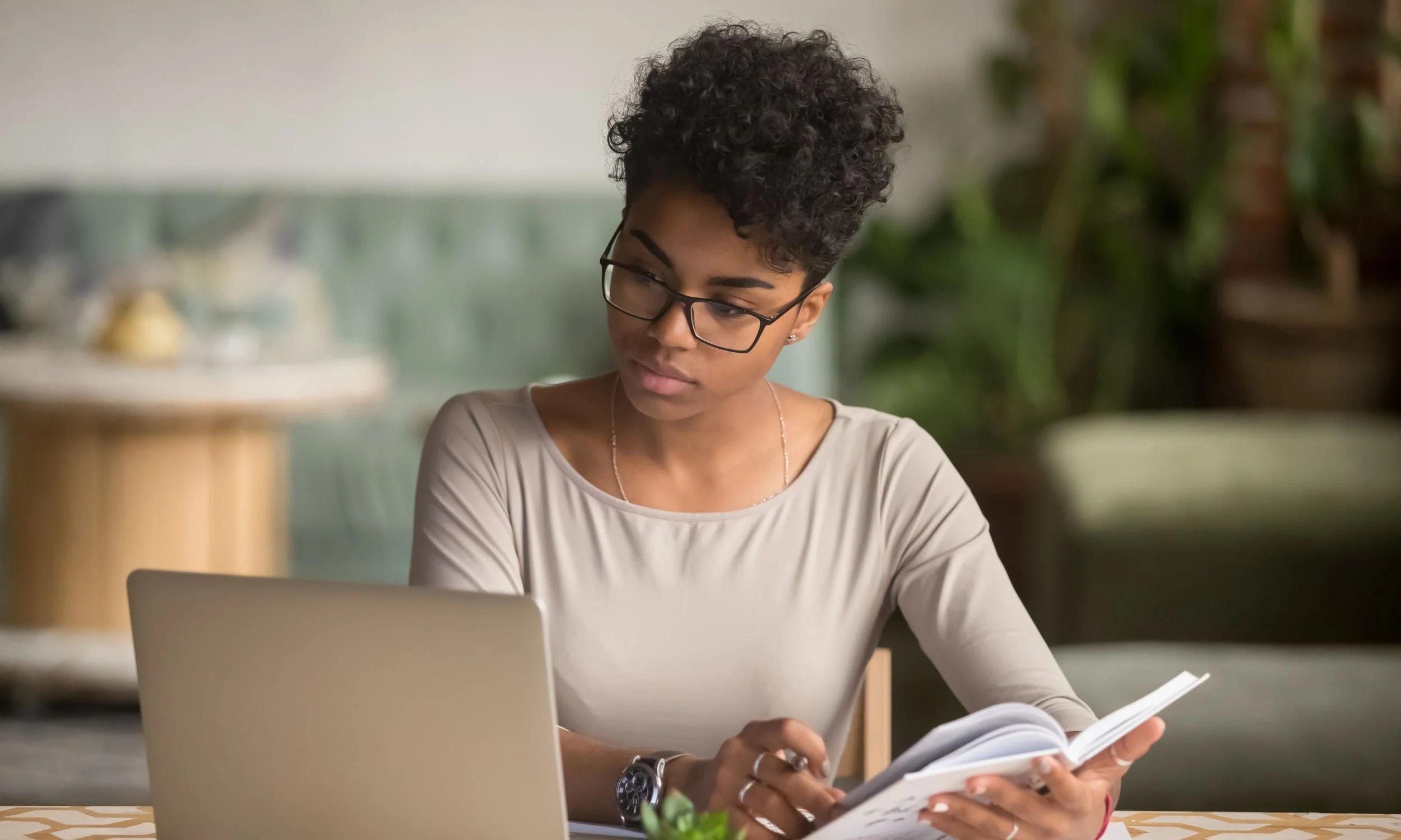 A woman looking at a laptop while holding an open book in one hand and a pen in the other.