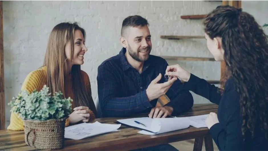 A woman hands keys to a smiling couple seated at a table with paperwork.