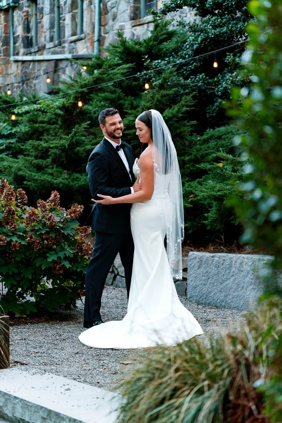 A bride and groom sharing a kiss outdoors, surrounded by green bushes and trees, with string lights overhead, during a wedding ceremony.