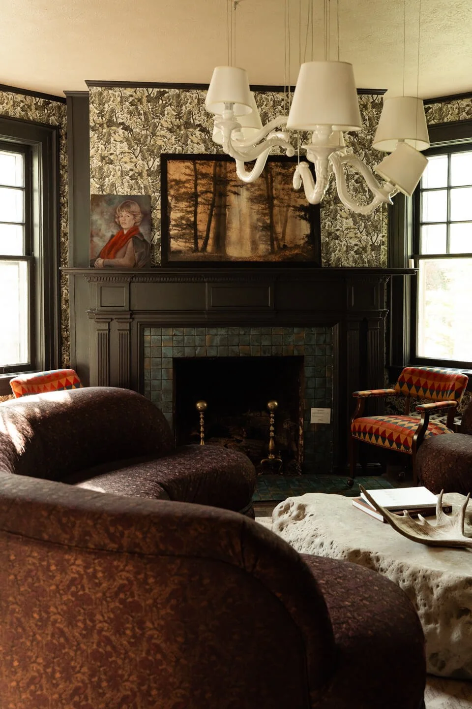 Living room with dark fireplace, floral wallpaper, colorful armchair, velvet sofa, artwork, antler decor, and large windows.