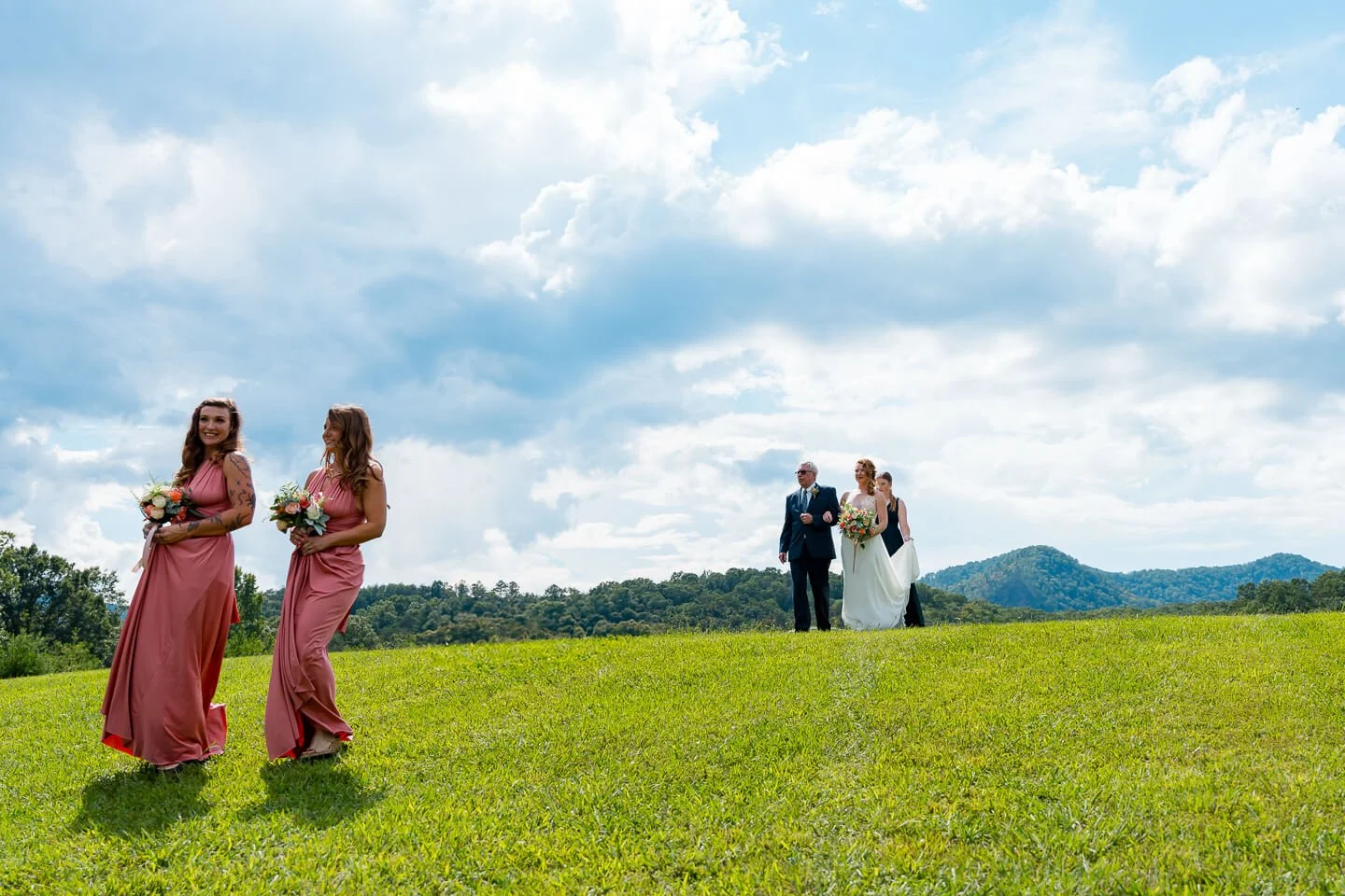 Wedding procession walking across a grassy field with scenic mountains and cloudy sky in the background.