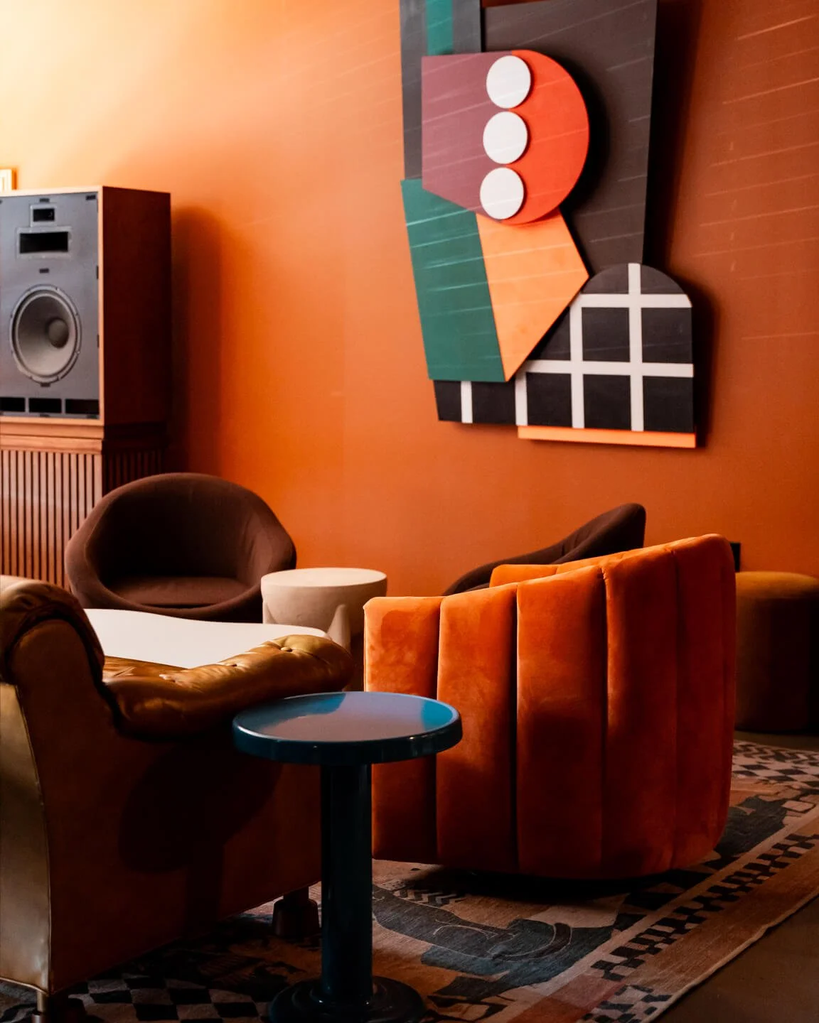 Interior of a stylish lounge with orange walls, modern art on the wall, a variety of colorful armchairs, a small round table, a speaker, and a patterned carpet.