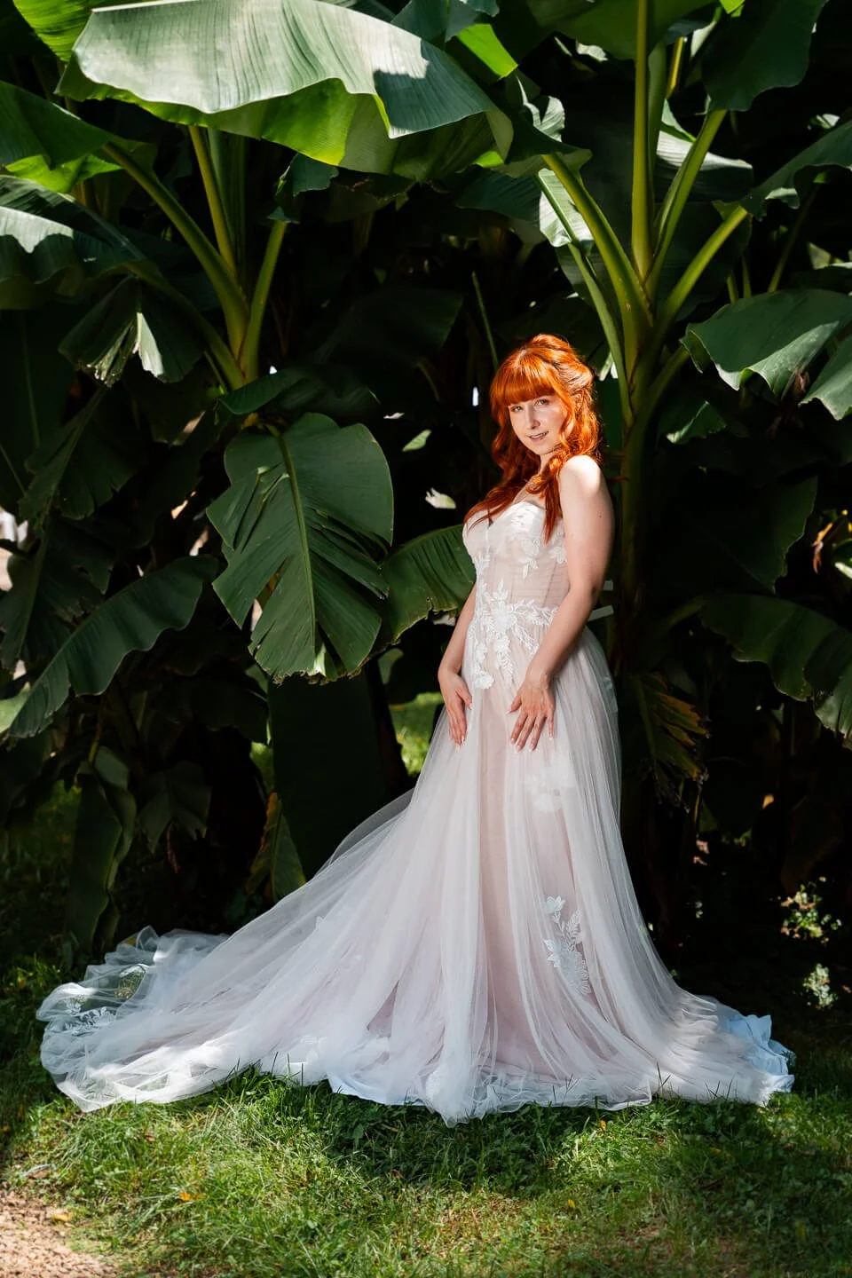 A woman with red hair in a white wedding dress standing outdoors among large green tropical leaves.