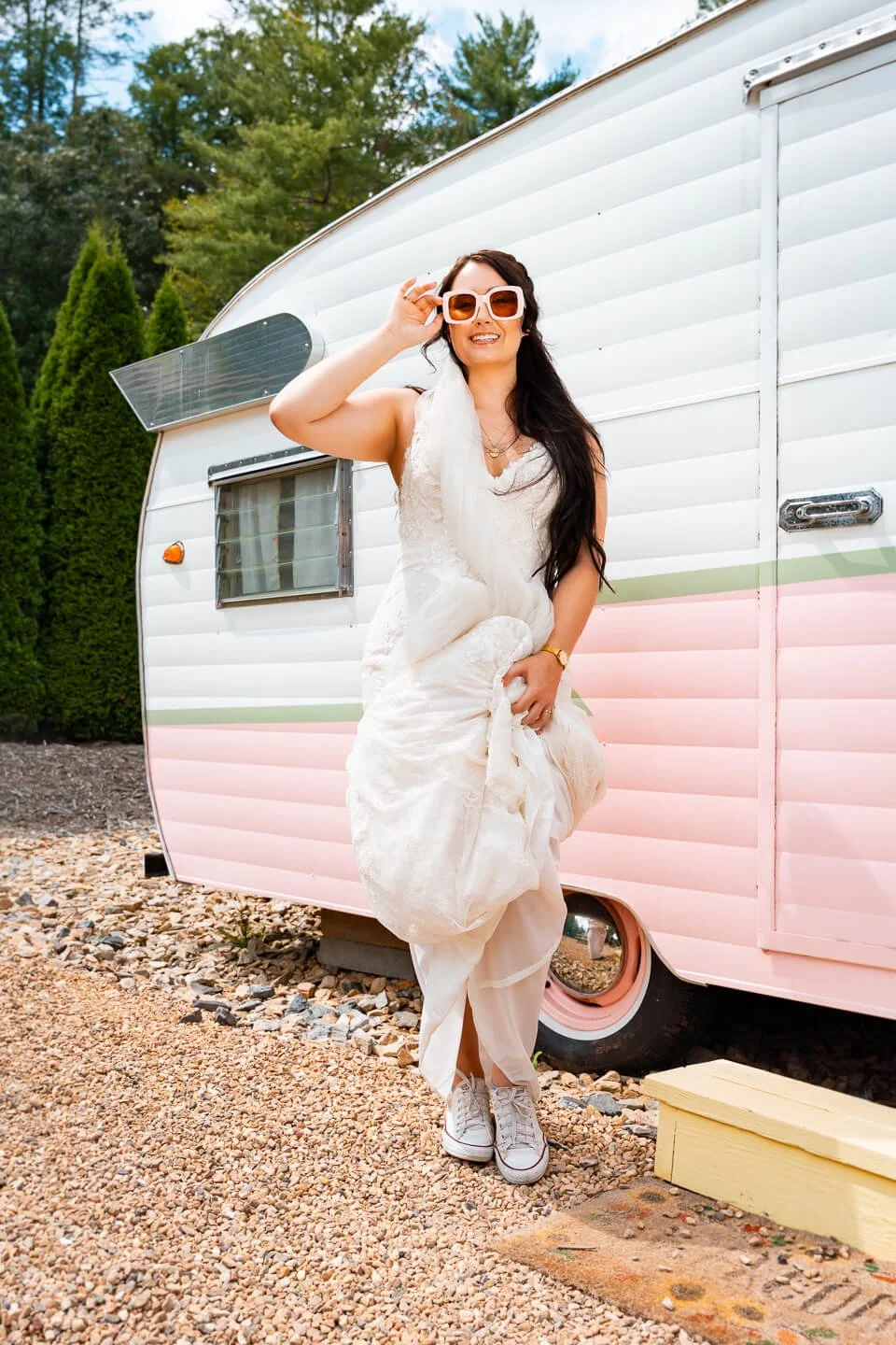 Woman in a white dress and sunglasses standing next to a vintage pink and white camper trailer outdoors.