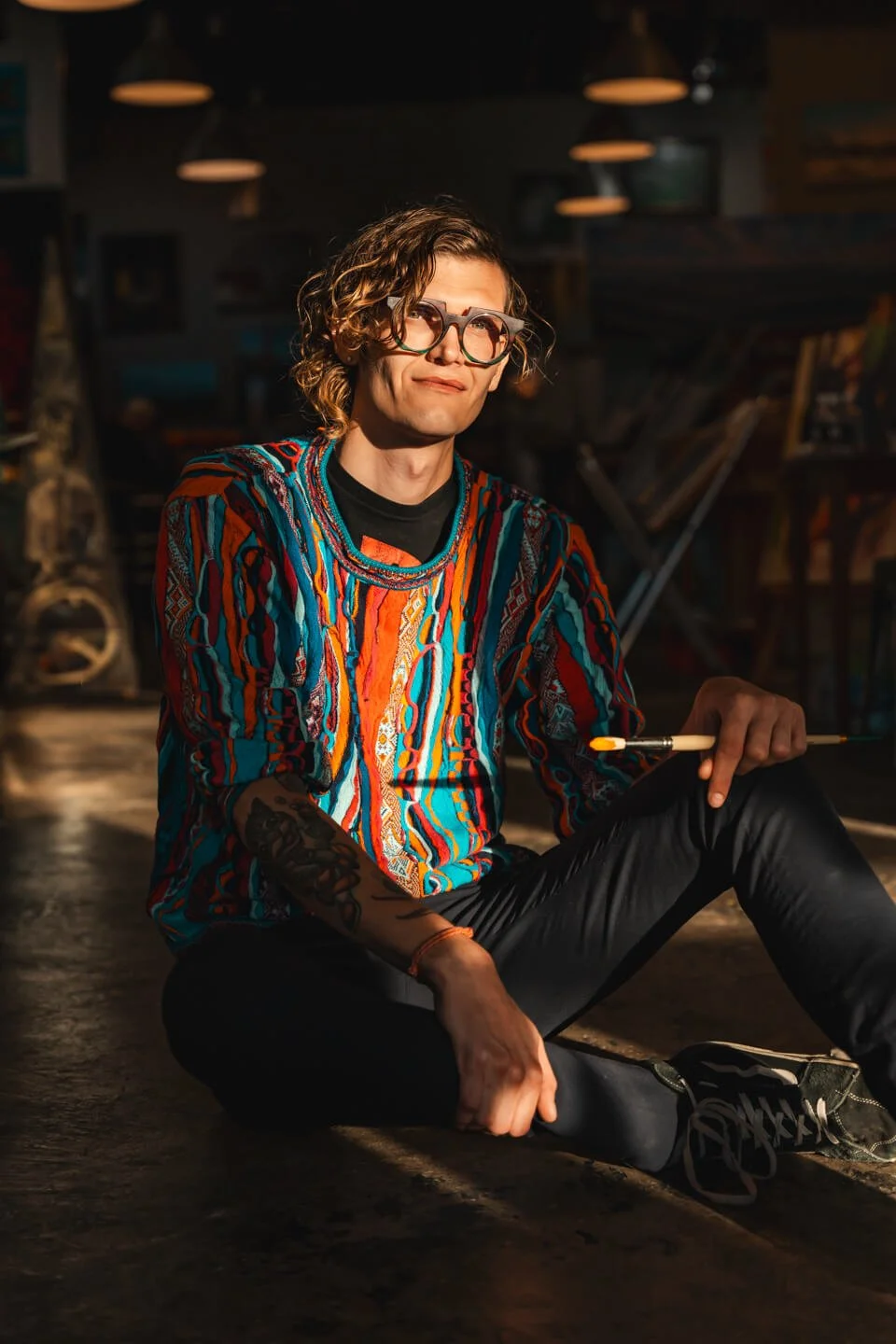 A person with curly hair, glasses, and tattoos sitting on the floor in a dimly lit art studio, holding a paintbrush.