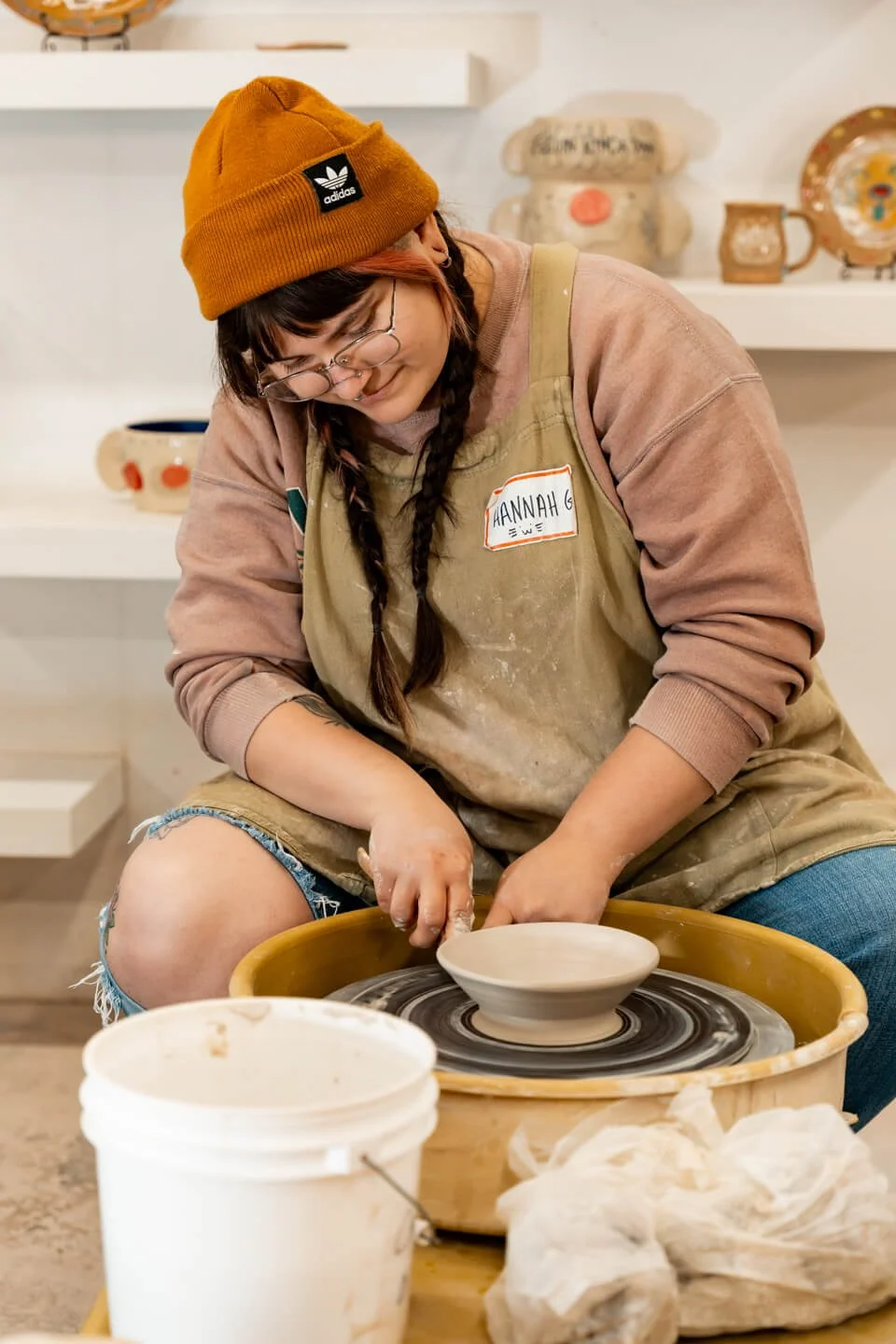A young woman with glasses, braided hair, wearing a brown beanie and a beige apron, is sculpting a ceramic bowl on a pottery wheel. She appears focused, working on the clay creation.