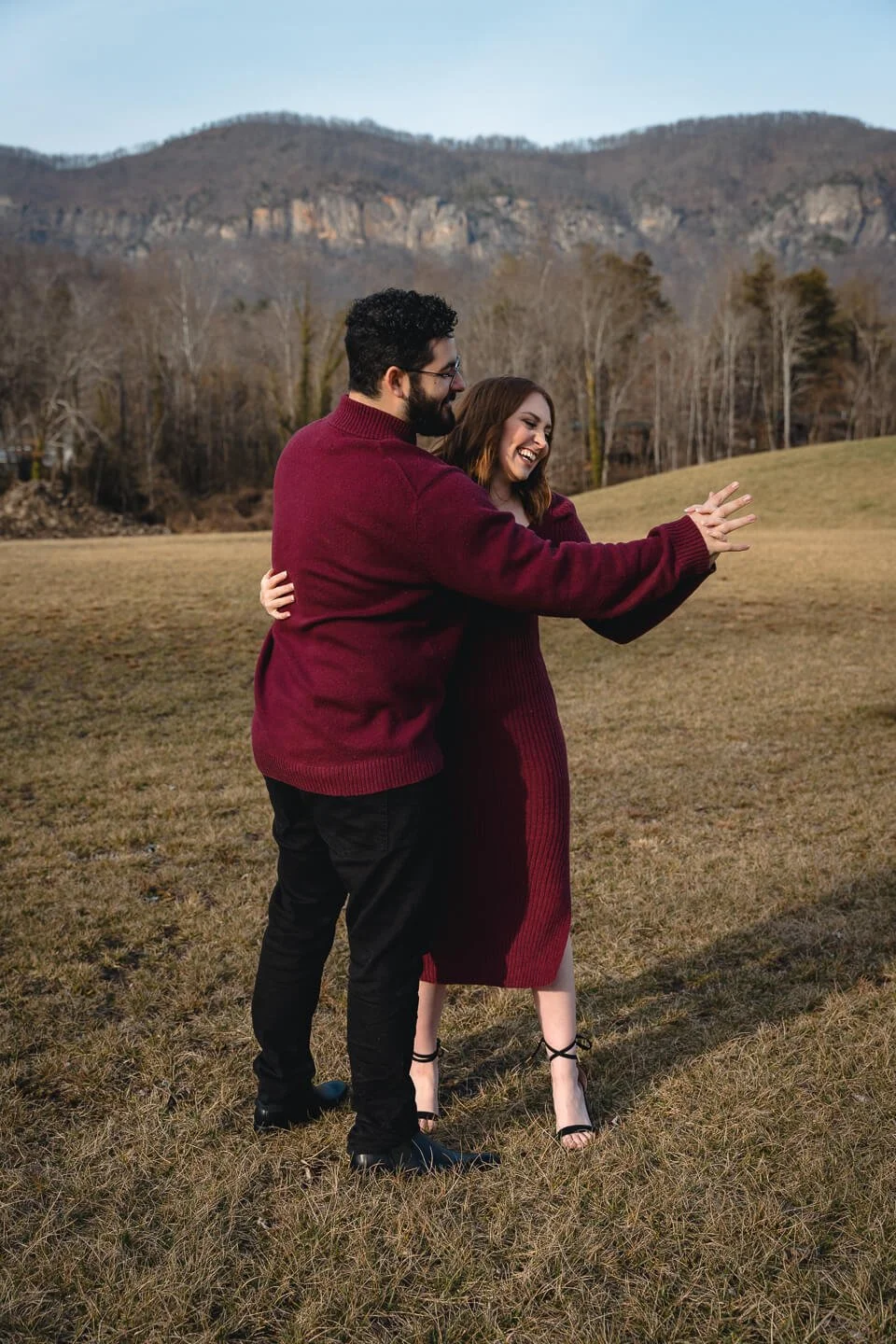 A couple standing in an open field with mountains in the background, smiling and taking a selfie together.