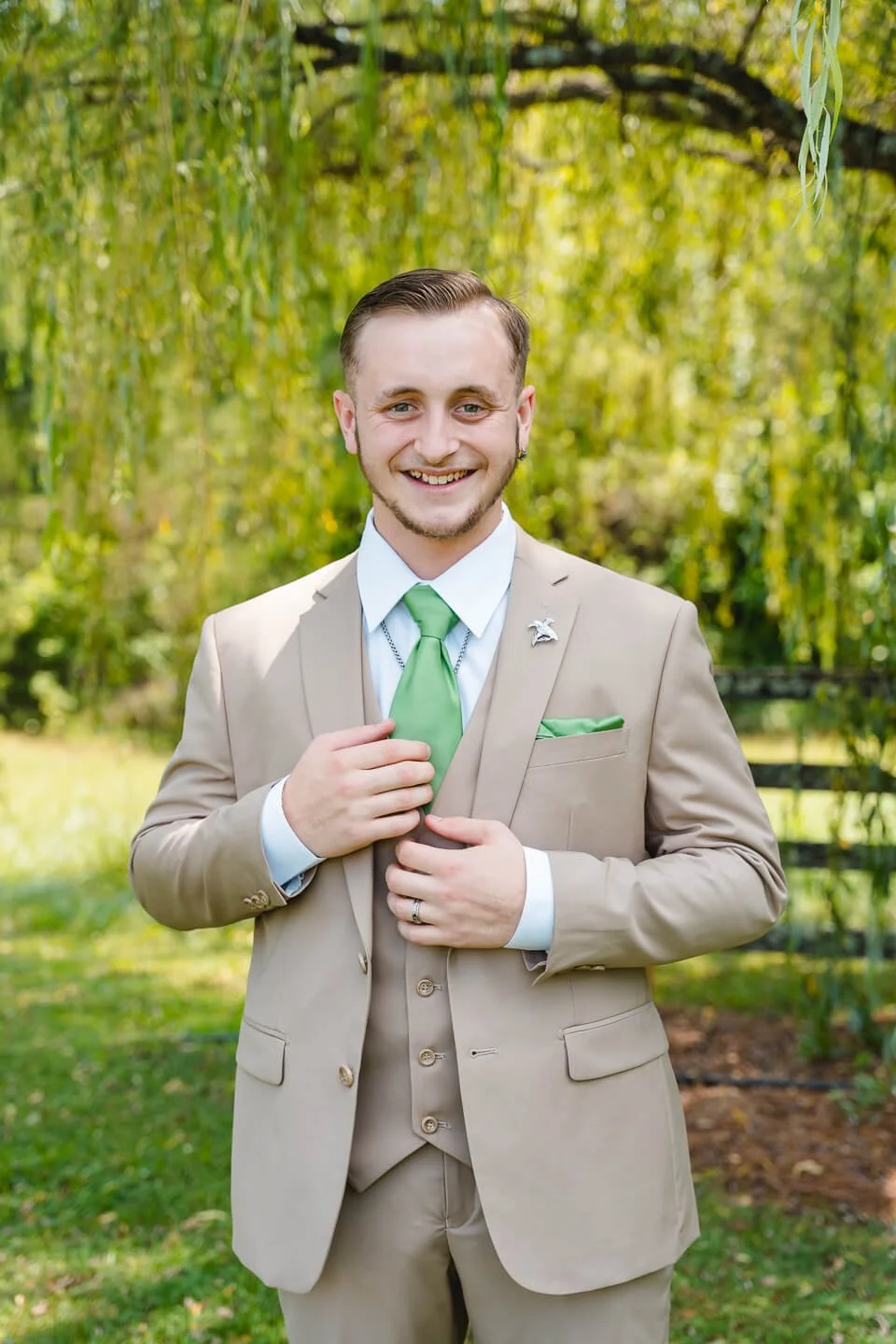 A smiling man in a beige suit, white shirt, and green tie standing outdoors in front of lush green trees, holding his tie with one hand and adjusting his suit jacket with the other.