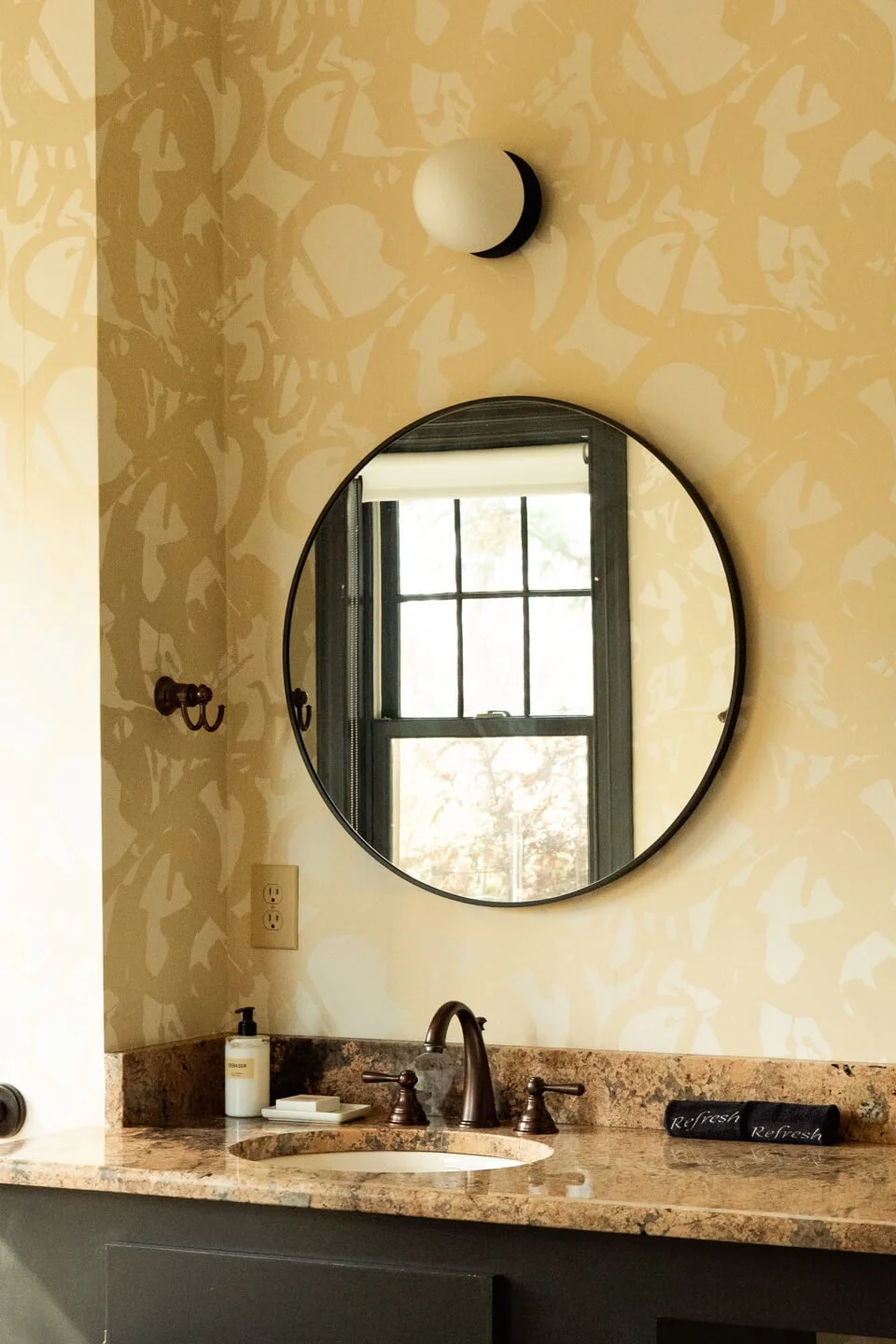 A bathroom vanity with a beige and brown speckled granite countertop, a bronze faucet, a soap dispenser, a mirror reflecting a window, and a black towel with the word "Refresh" written on it. The wall behind has a beige pattern and is lit by a round
