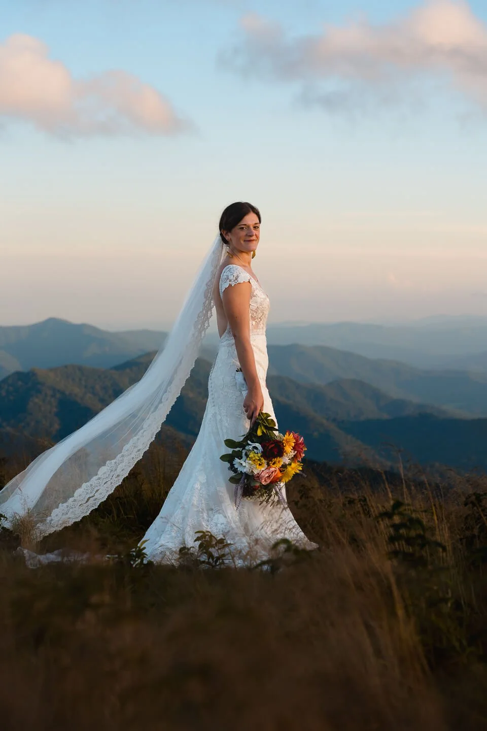 A bride in a white lace wedding dress holding a bouquet of colorful flowers standing on a hill with mountain views in the background during sunset.