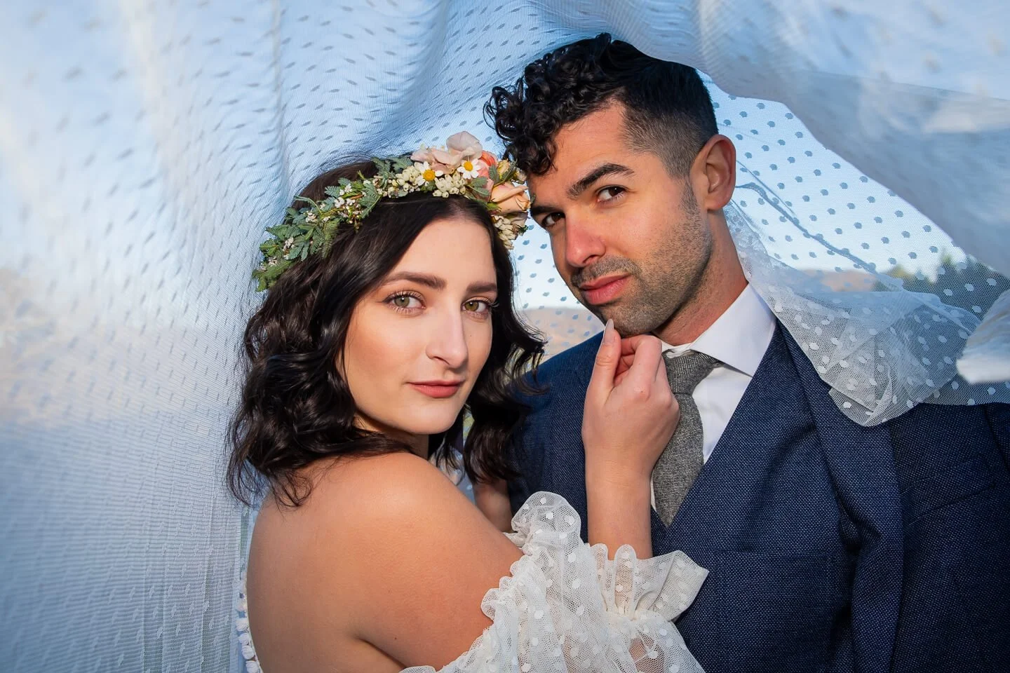 A newlywed couple in wedding attire, with the woman wearing a floral crown and the man in a suit, posing closely under a white cloth canopy.