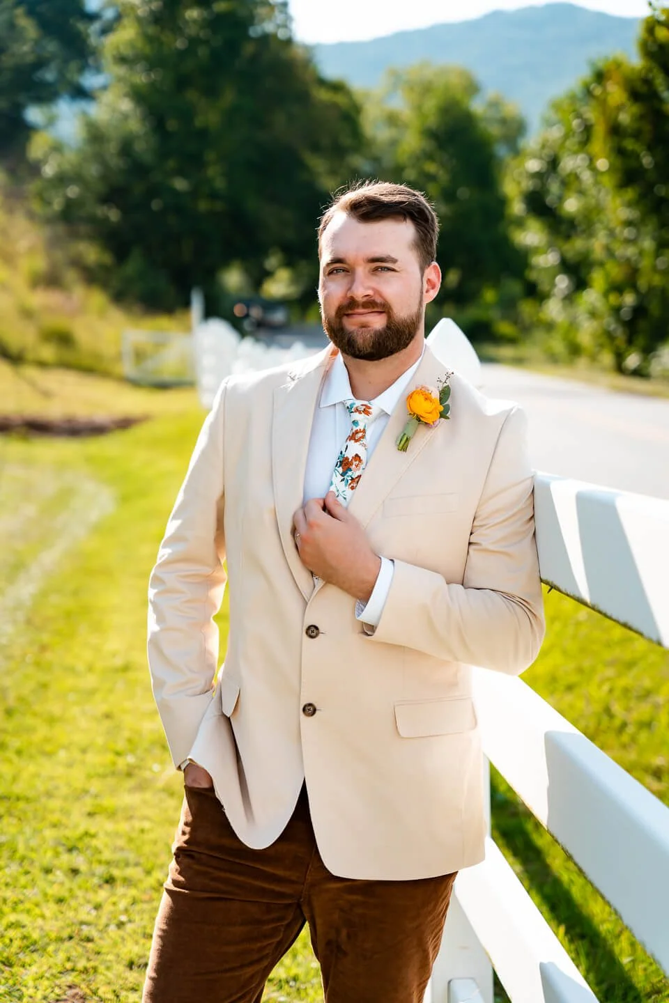 A man in a cream-colored blazer and brown pants, with a floral tie and a yellow boutonniere, leaning against a white fence outdoors during daytime, with trees and mountains in the background.