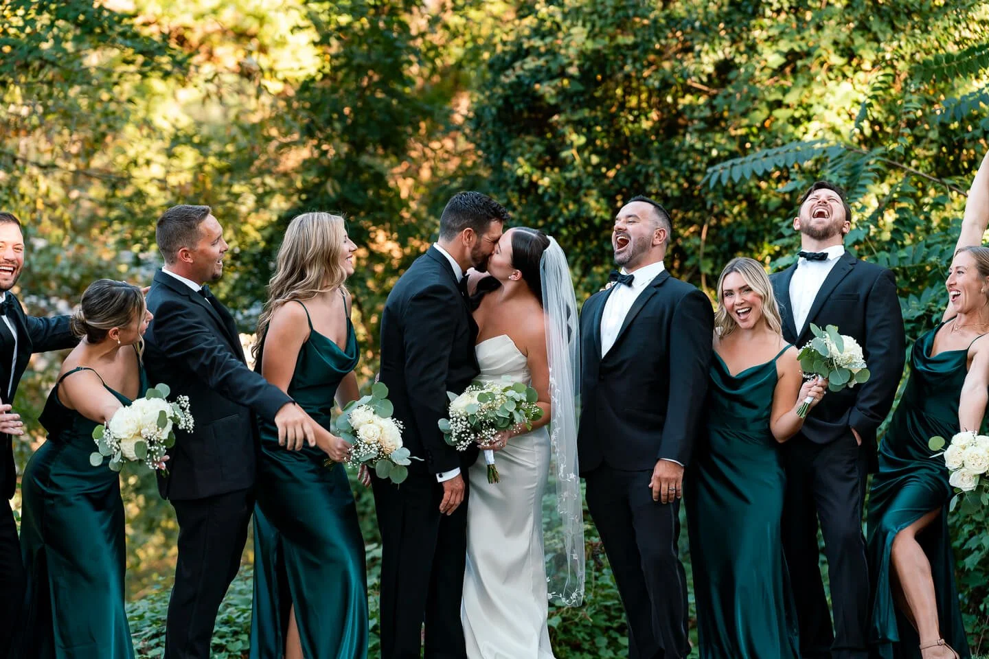A wedding celebration with the bride and groom kissing, surrounded by their wedding party in formal attire outdoors, smiling and laughing.