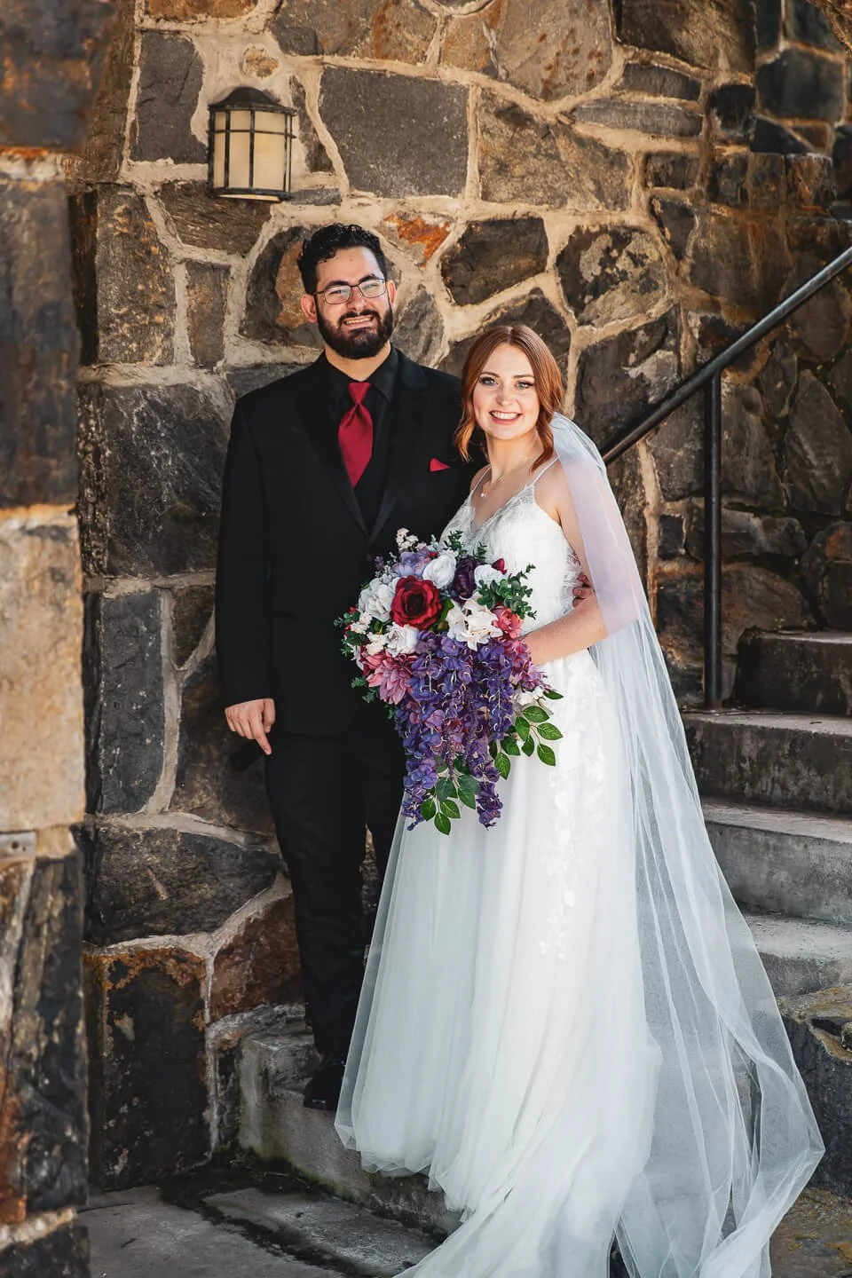 A wedding couple standing beside a stone staircase and wall. The bride is holding a large, colorful bouquet and wearing a white wedding dress with a long veil. The groom is wearing a black suit with a red tie and pocket square. They are smiling and p