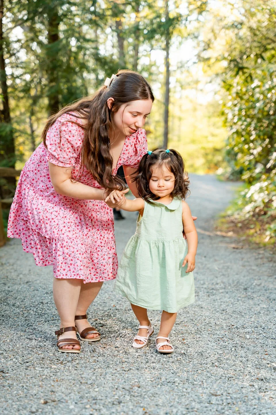 A woman wearing a pink floral dress is helping a young girl in a light green dress walk on a gravel path in a wooded area.
