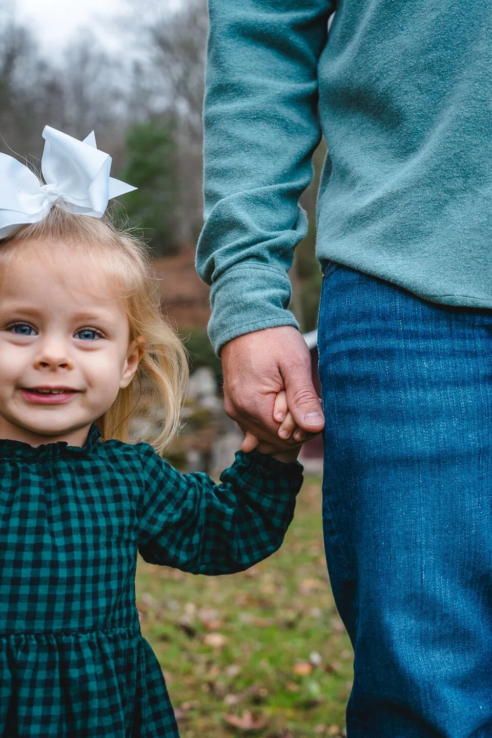 A young girl with blonde hair wearing a green and black checked dress and a white bow in her hair, holding an adult's hand outdoors.