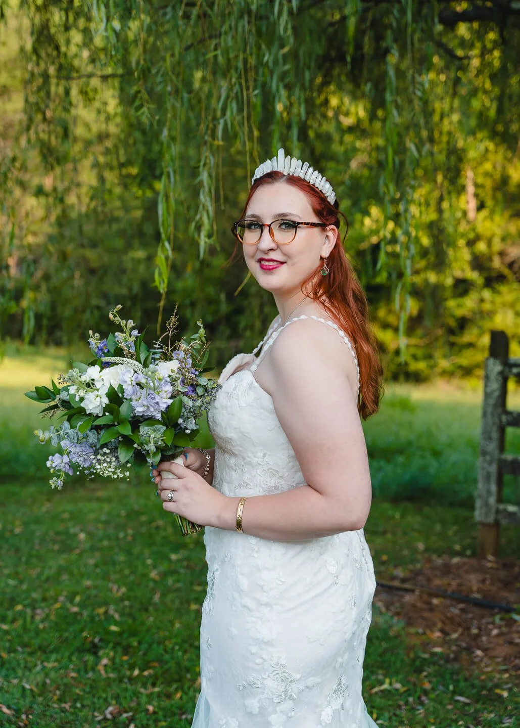 A bride with red hair in a white lace wedding gown holding a bouquet of flowers outdoors, smiling, with green foliage and hanging branches in the background.