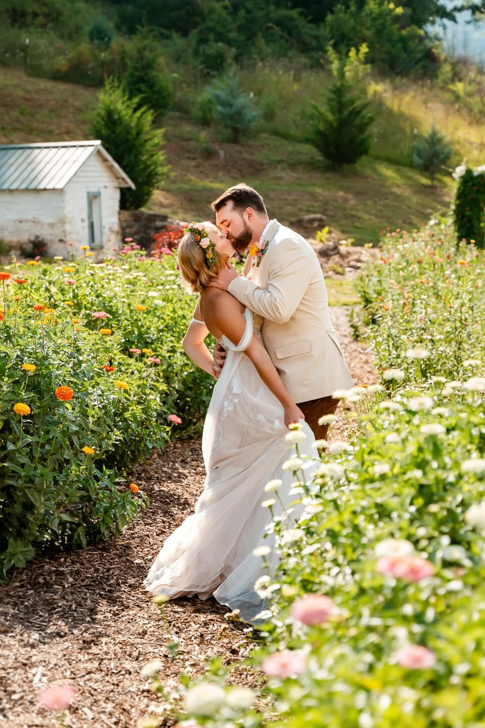 A bride and groom sharing a kiss in a lush garden filled with blooming flowers, with a small white shed and green hills in the background.