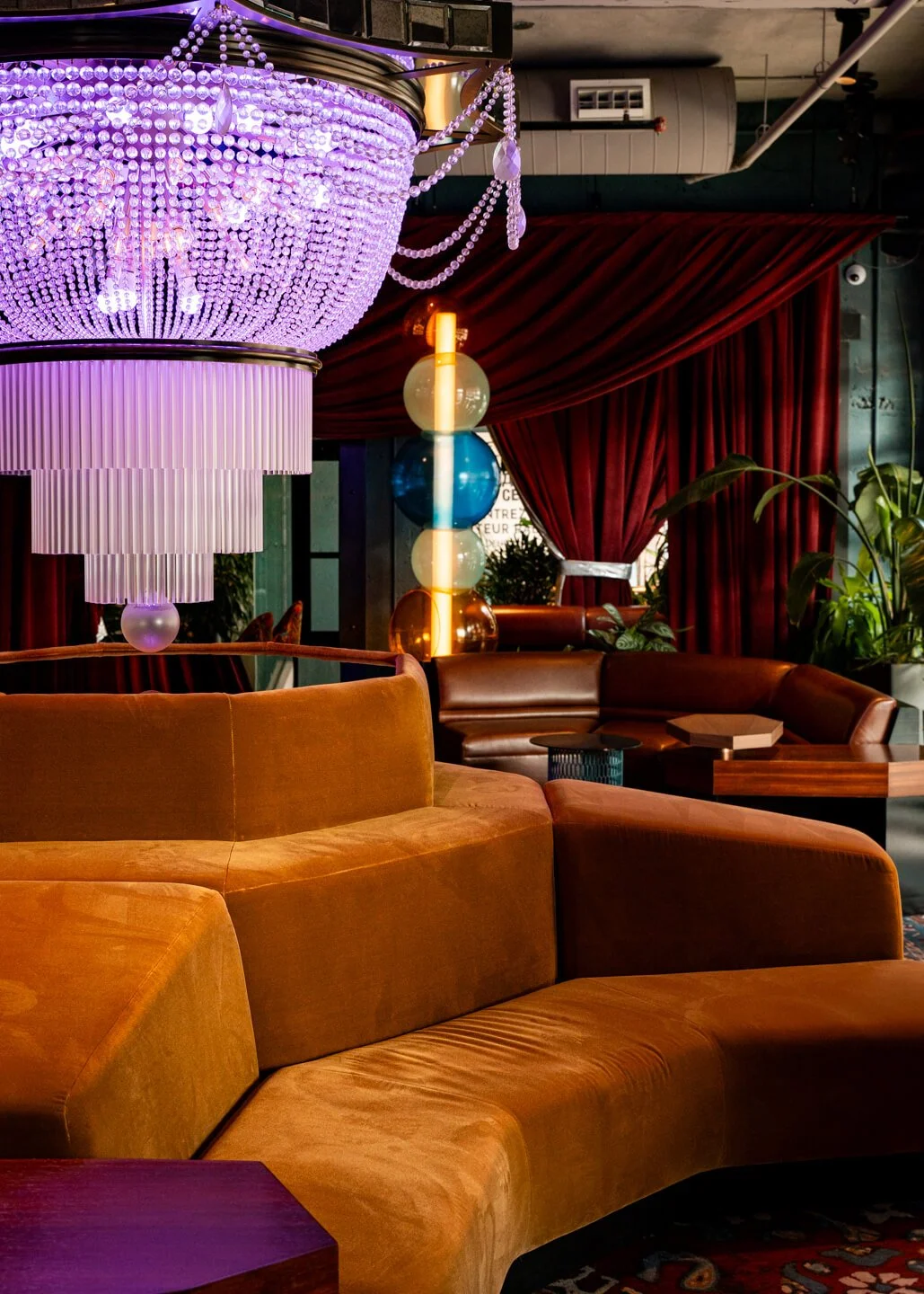 Modern lounge with a large, purple chandelier, red curtains, brown leather sofas, and decorative balloon sculpture with blue, white, orange, and gold balloons.