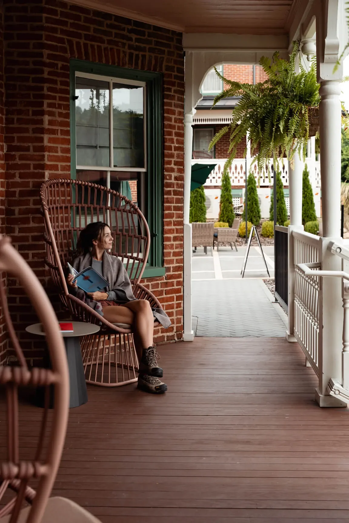 Woman sitting on a porch in a hanging chair, reading a book, with a small round table nearby, and a view of a garden and outdoor seating area in the background.