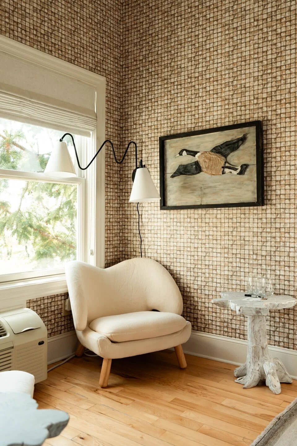 A cozy corner with a white armchair, a small abstract side table, and a wall-mounted lamp next to a window with greenery outside. There is framed artwork of a horse on the wall, and a patterned wallpaper covers the walls.