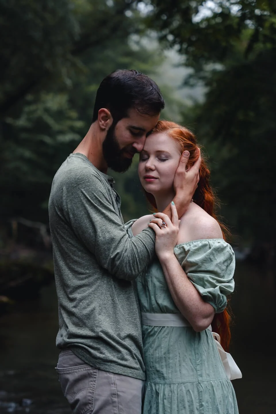 A man and woman embrace tenderly outdoors, with the man gently touching the woman's face and forehead as they close their eyes, surrounded by lush greenery.