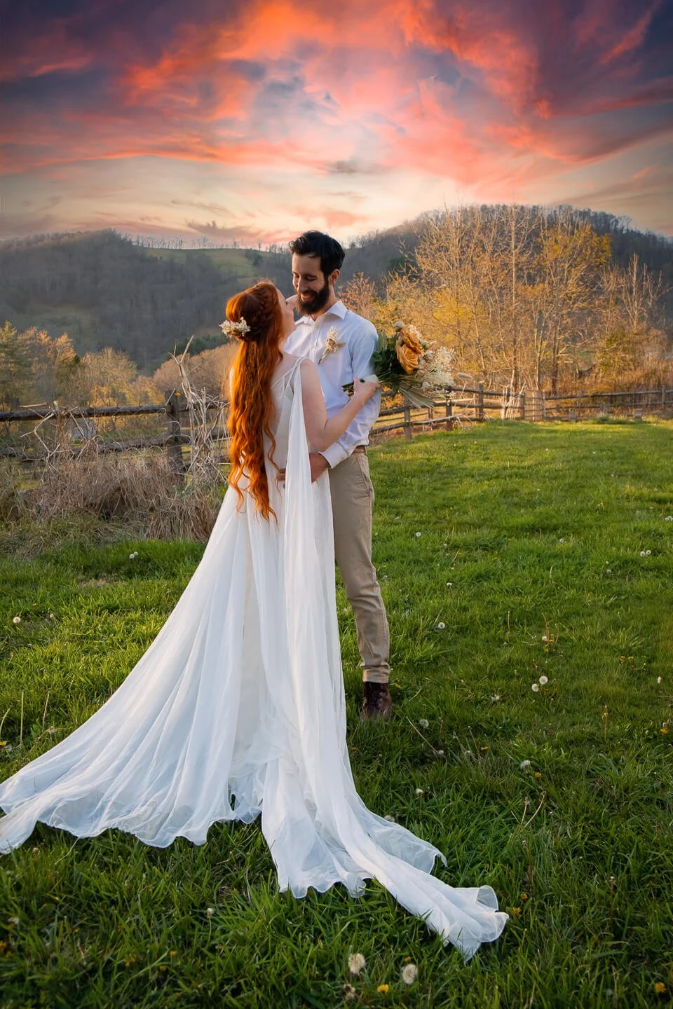 A newly married couple stands in a grassy field during sunset, with the woman in a flowing white wedding gown and the man in a white shirt and khakis. They are smiling at each other, with the woman holding a bouquet of flowers, and the sky is filled