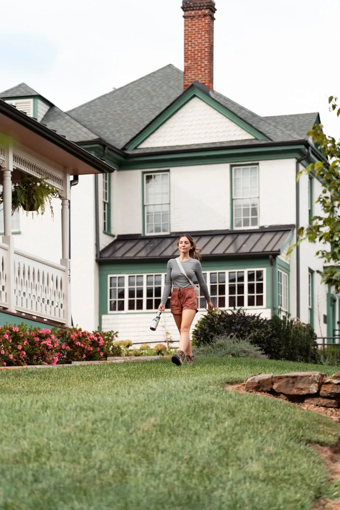 A woman walking on a well-manicured lawn in front of a large, vintage-style house with green trim and a brick chimney. She is holding a small bottle in her right hand and has a crossbody bag.