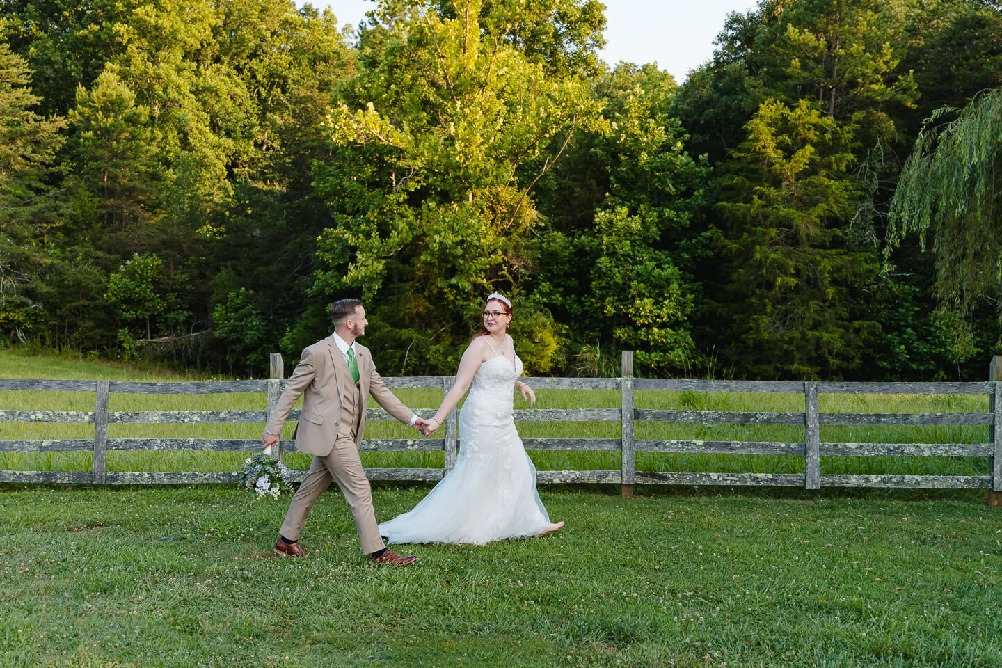 A bride and groom holding hands walking outdoors on grass, with a wooden fence and lush green trees in the background.