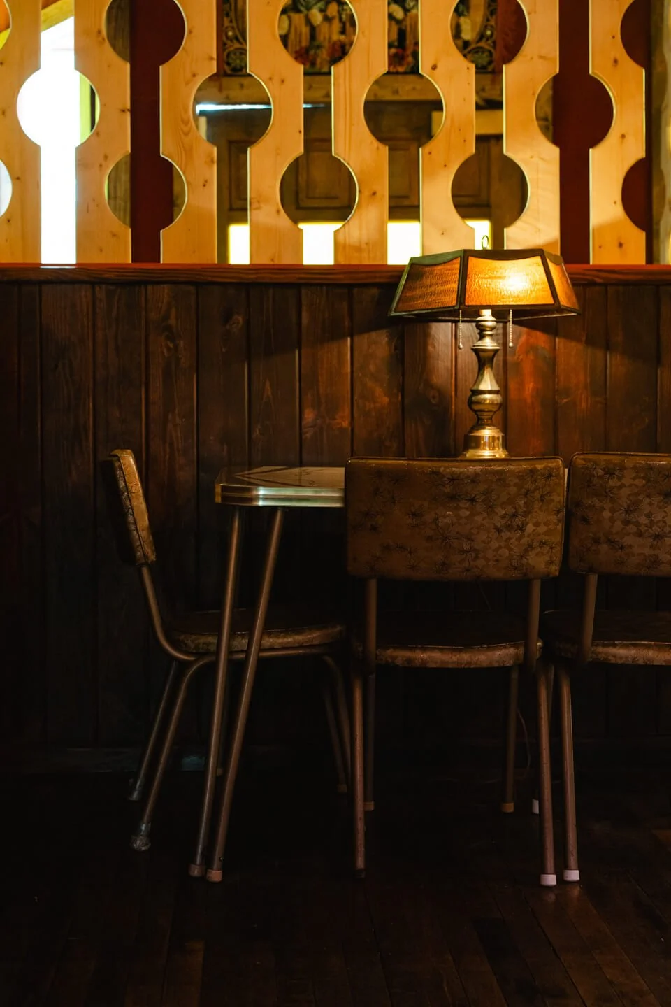 A dimly lit cozy corner with a small round table, two vintage chairs with floral upholstery, a vintage table lamp with a warm glow, and a decorative wooden partition with circular cutouts in the background.