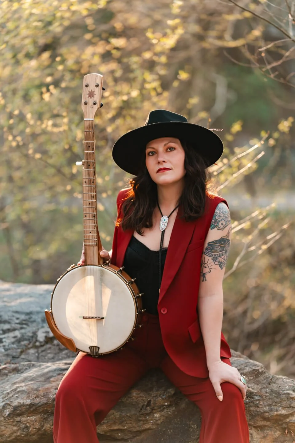 A woman wearing a black wide-brim hat and a red sleeveless suit seated outdoors on a rock, holding a vintage banjo, with a background of trees and autumn foliage.