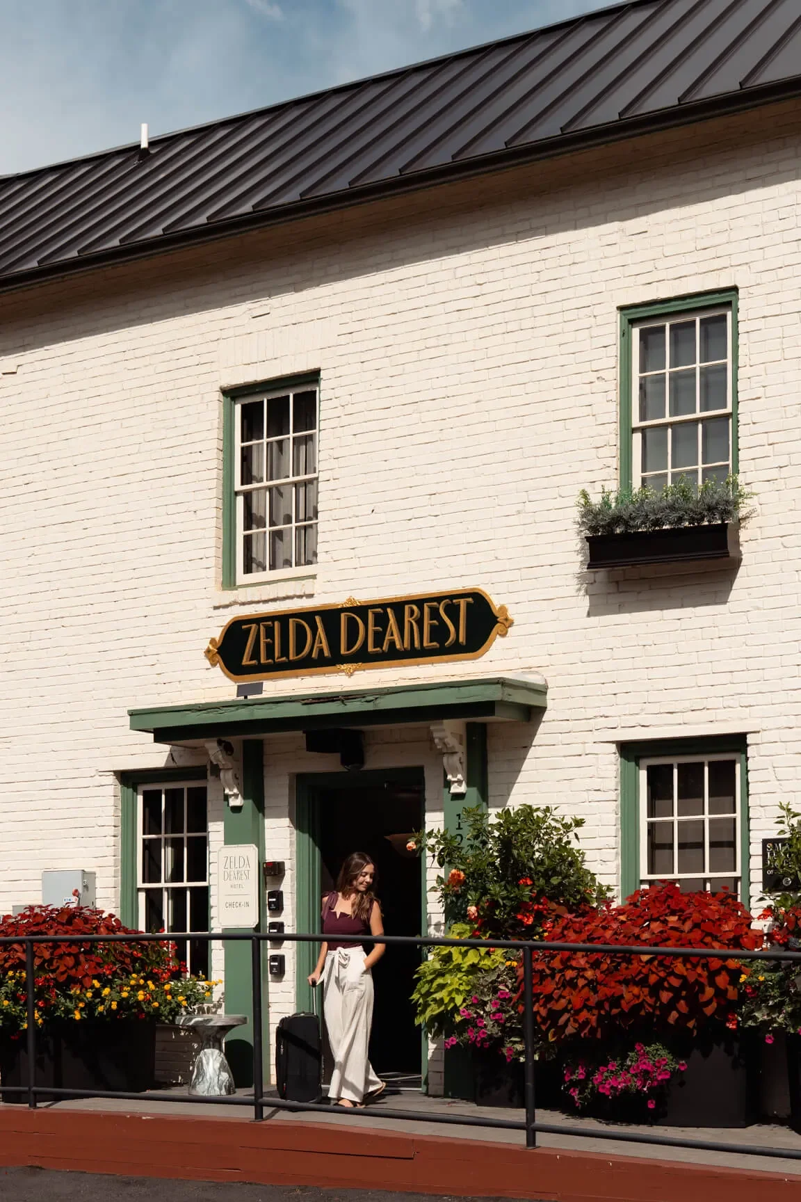 A woman standing at the entrance of the Zelda Dearest hotel, with a suitcase, surrounded by colorful flowers, a white brick building, and a sign that reads 'Zelda Dearest' above the doorway.