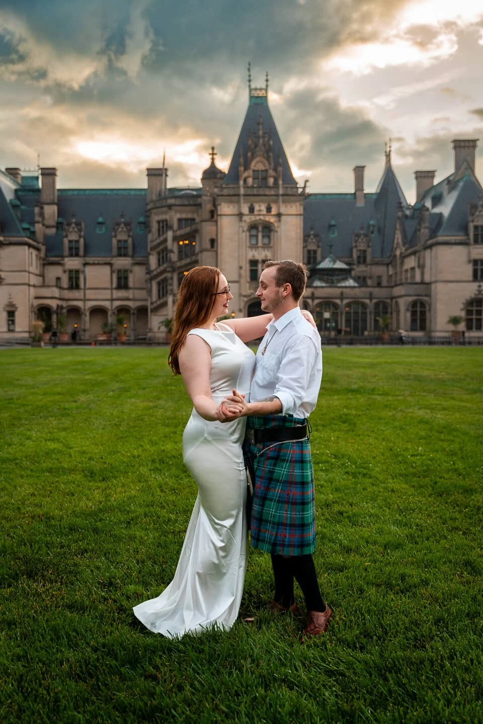A couple dancing outdoors on green grass in front of a large castle with tall spires and turrets during sunset.