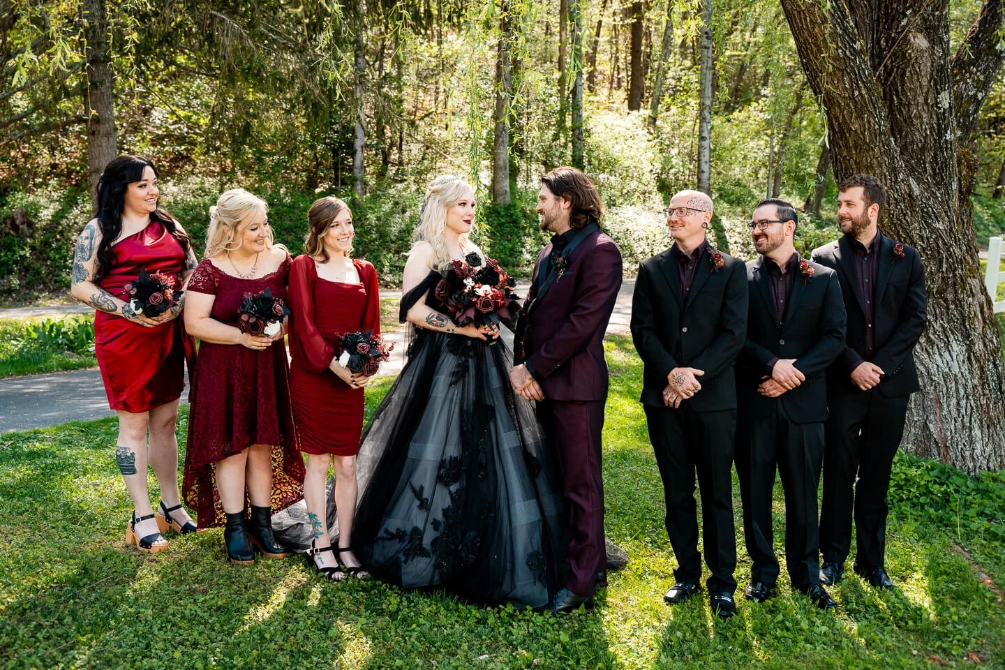A wedding party in a wooded outdoor setting with the bride and groom holding hands, surrounded by bridesmaids and groomsmen dressed in elegant formal attire, with all members smiling and looking at each other.